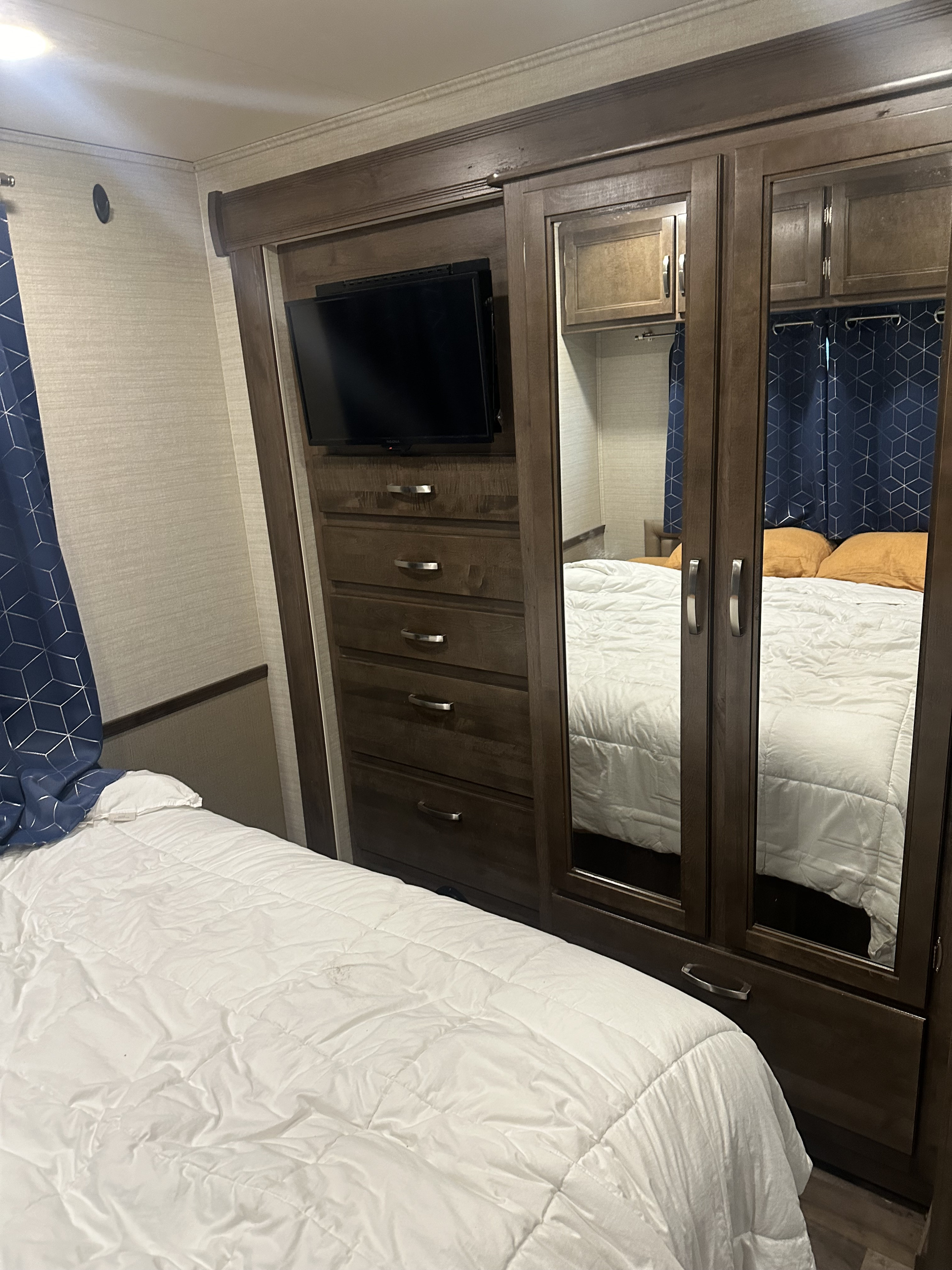 Cozy RV rental bedroom showing a white bed, built-in storage with TV & drawers, and mirrored closet reflecting blue curtains.. Jayco Alante 2020