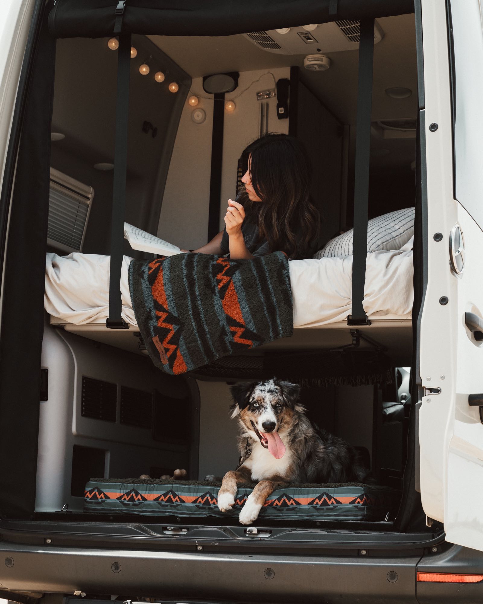 This cozy rv rental is pet-friendly! Woman reads in the upper bunk, dog relaxes on its bed in the van's open back.. Winnebago Revel 2022