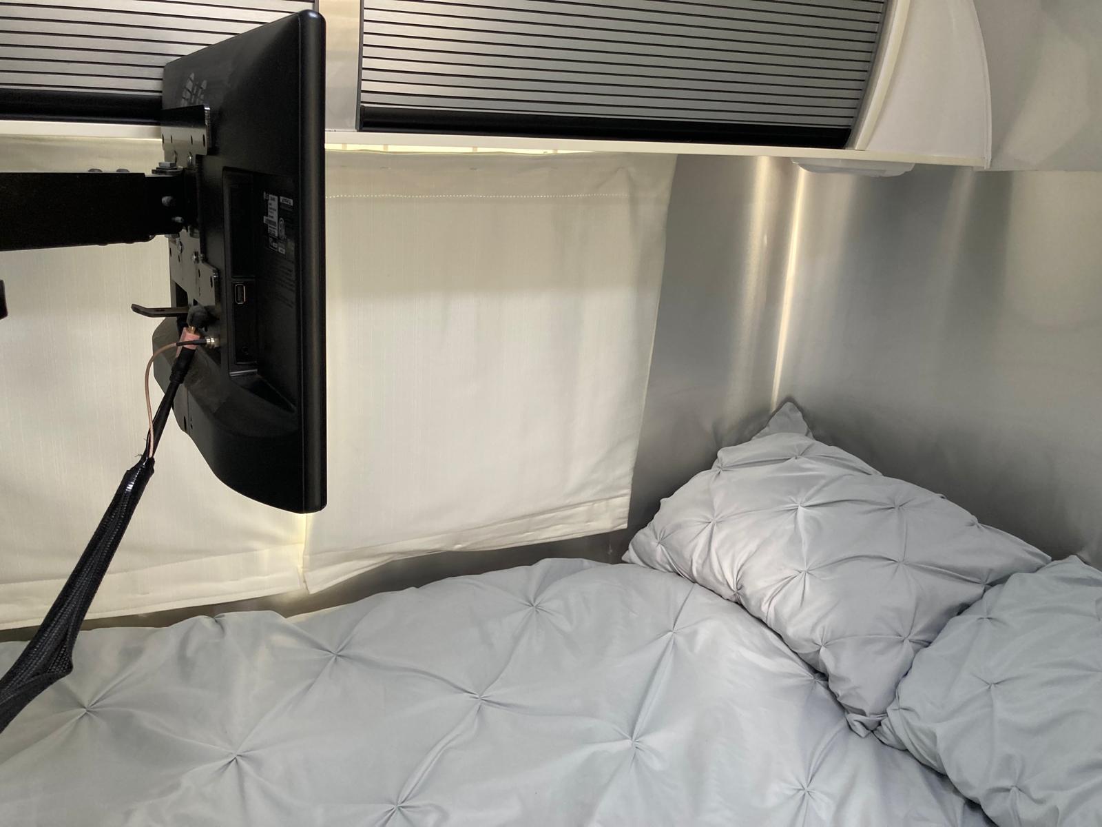Cozy RV rental bedroom with a mounted TV, soft grey quilted bedding, and metallic walls. Ideal for comfortable travel.. Airstream Sport 2017