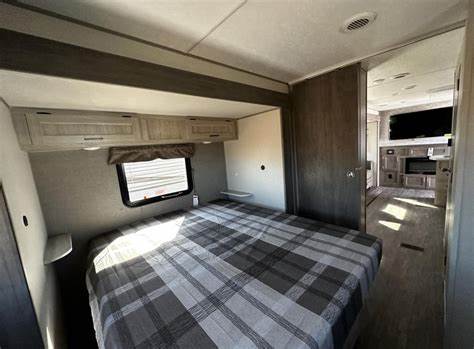 Modern RV rental interior: cozy bedroom with plaid bedding & spacious living area beyond.. Forest River Aurora 2024