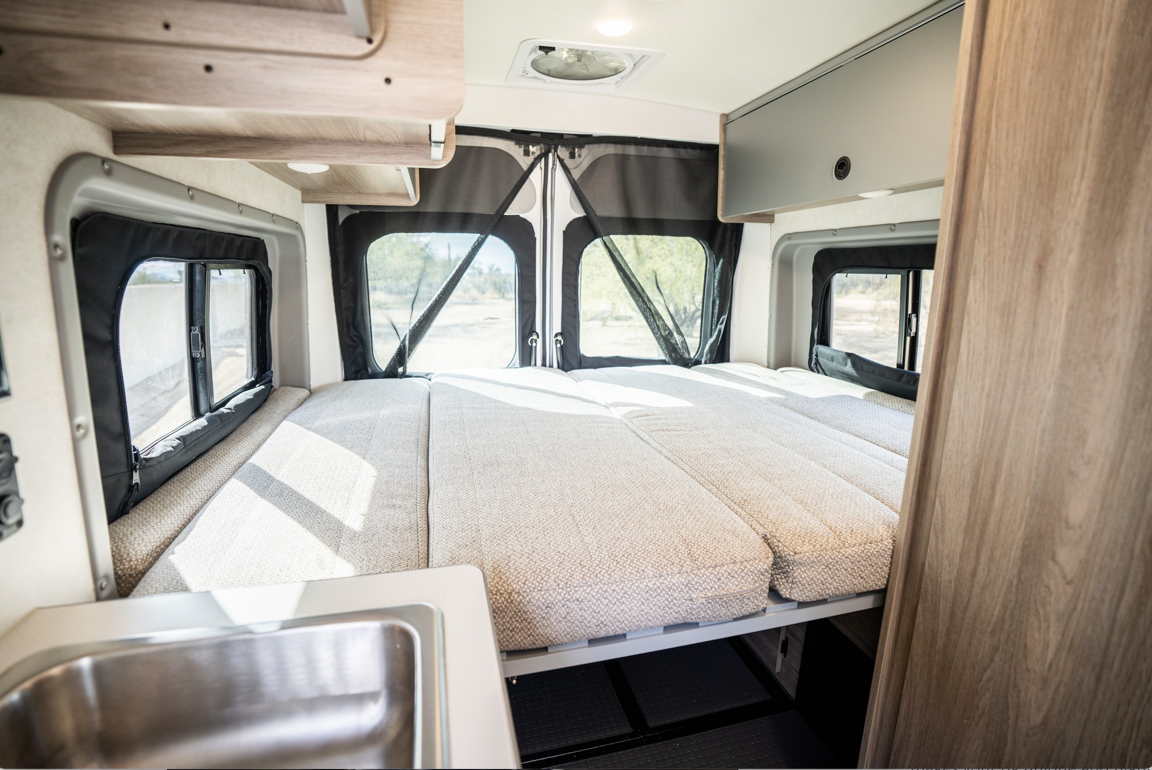 RV rental's functional interior: convertible bed, large windows revealing the outside, overhead storage, and a compact sink.. Winnebago Solis 2023