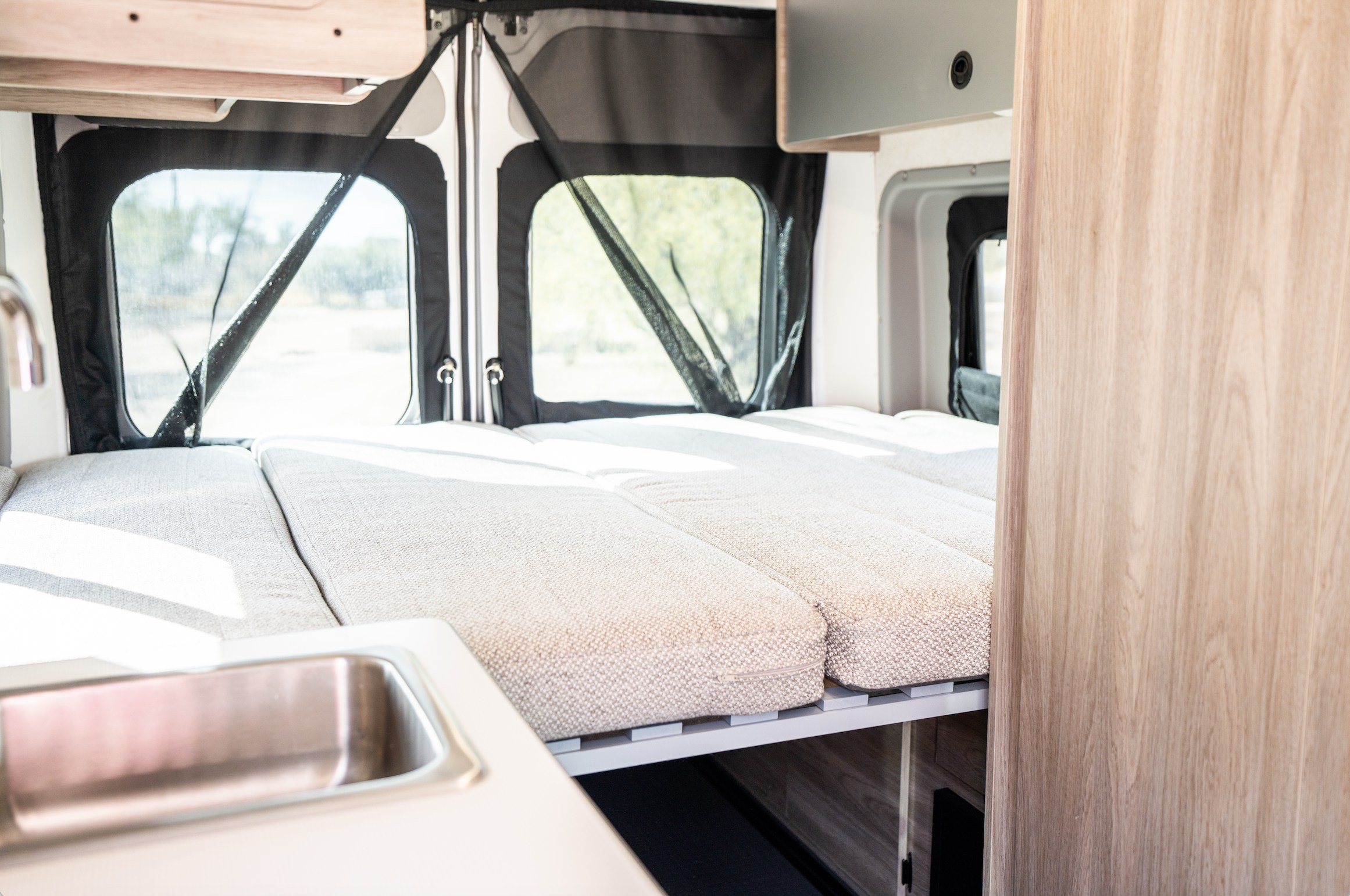 RV rental interior featuring a comfy bed with light cushions, large screened windows, and a small kitchen sink.. Winnebago Solis 2023