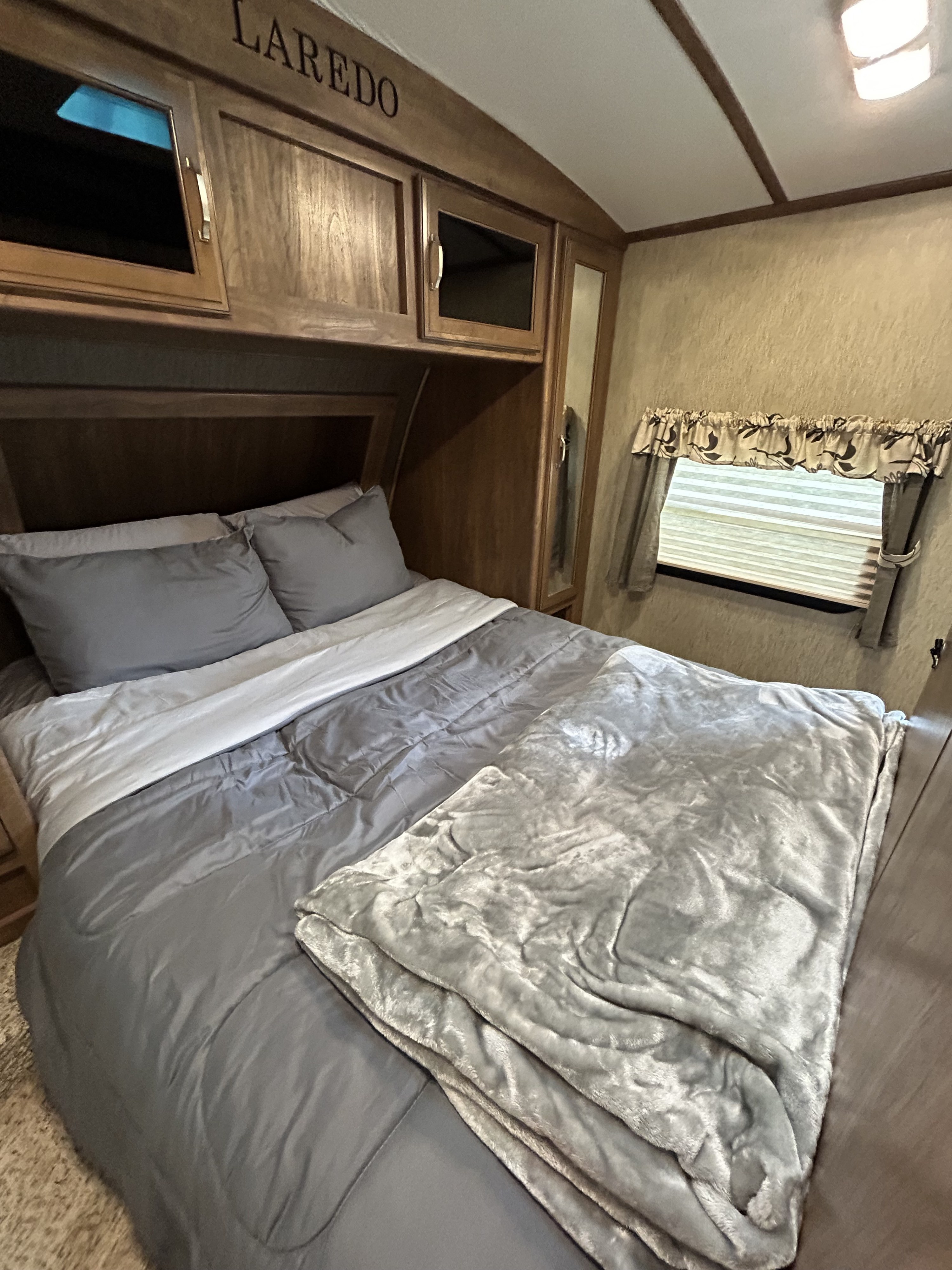 Inside an RV rental: a neat bedroom with gray bed linens, a plush throw blanket, wooden cabinets labeled Laredo, and window.. Keystone RV Laredo 2016
