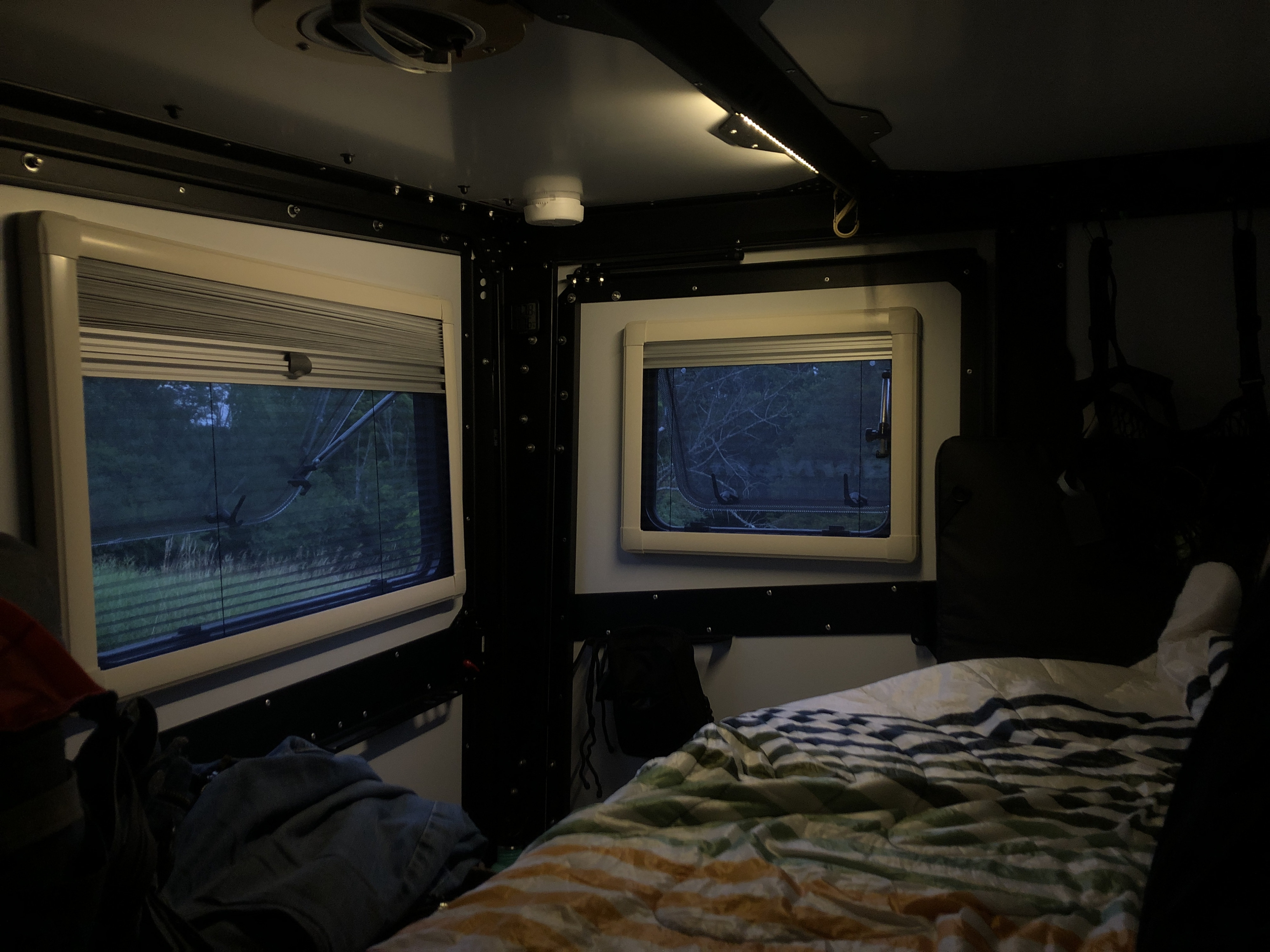 Cozy RV rental interior at dusk: two windows reveal a wooded view, complemented by a comfortable bed and soft ceiling lighting.. TAXA TAXA Trailer 2019