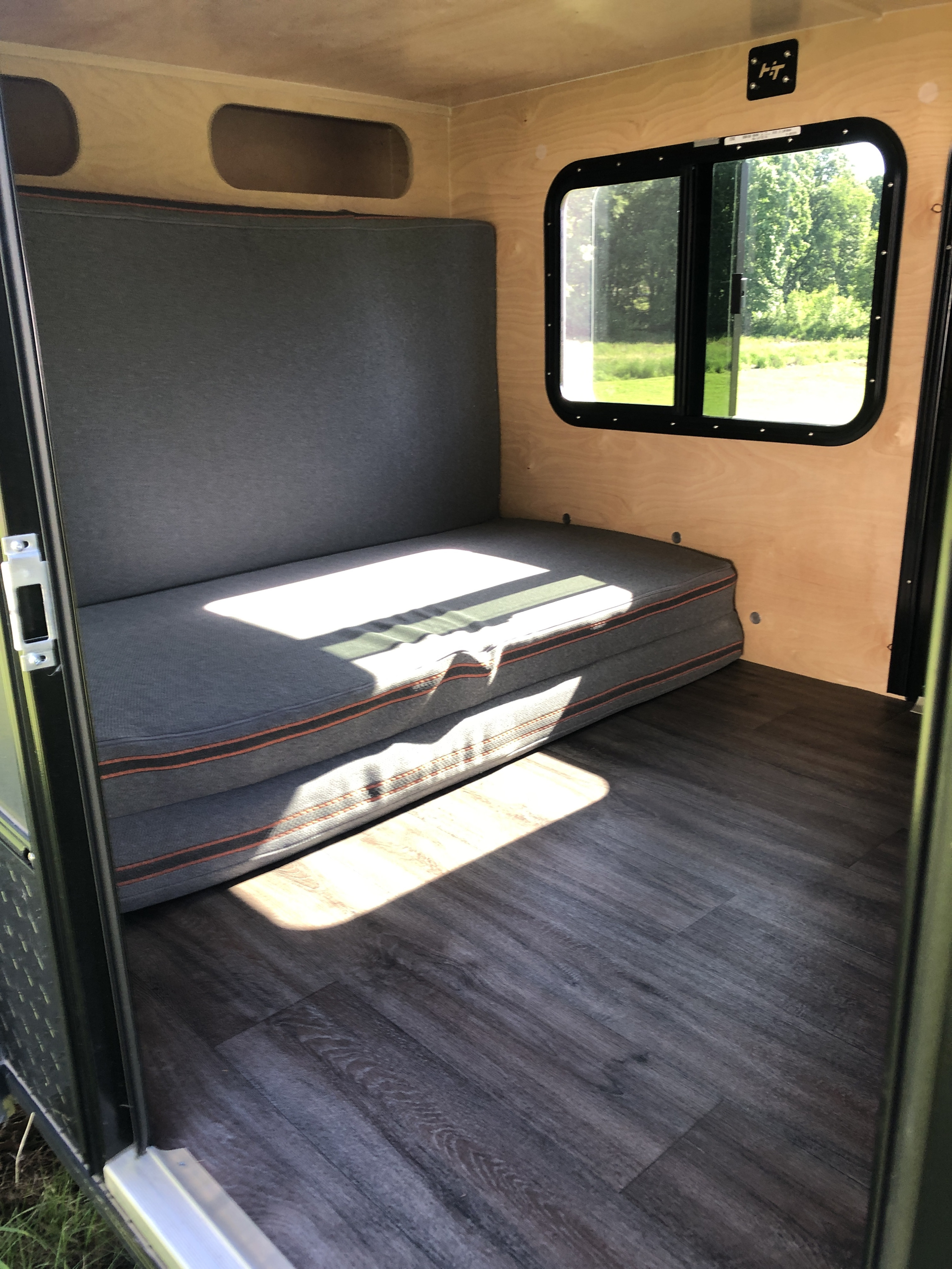 Cozy RV rental camper interior with wooden walls, dark gray seating/bed, wood-look flooring, and a bright window.. Hiker 5X9 MR 2023