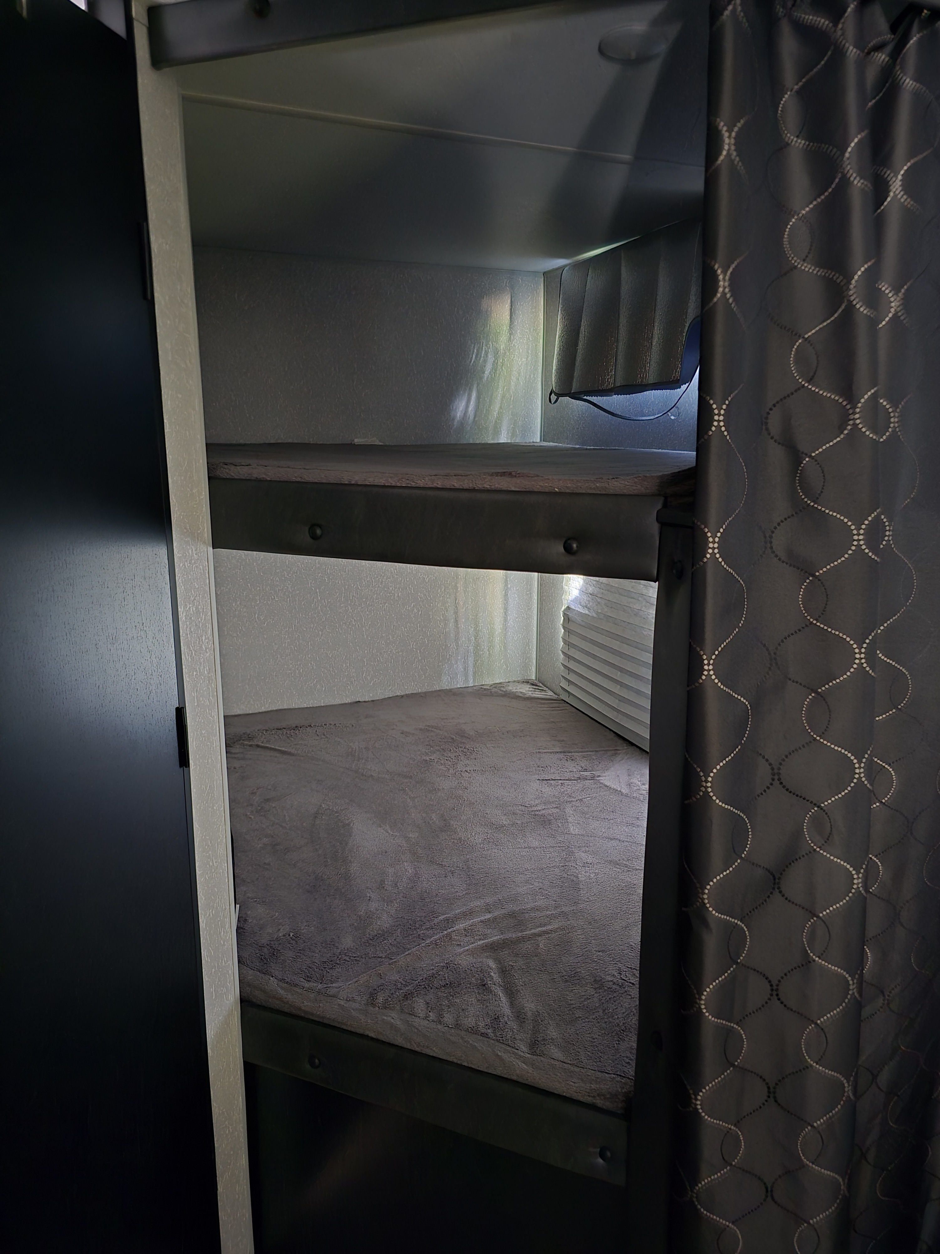 Interior view of an RV rental showing two compact bunk beds with grey bedding, framed by a dark patterned privacy curtain.. Forest River Cherokee Grey Wolf 2022