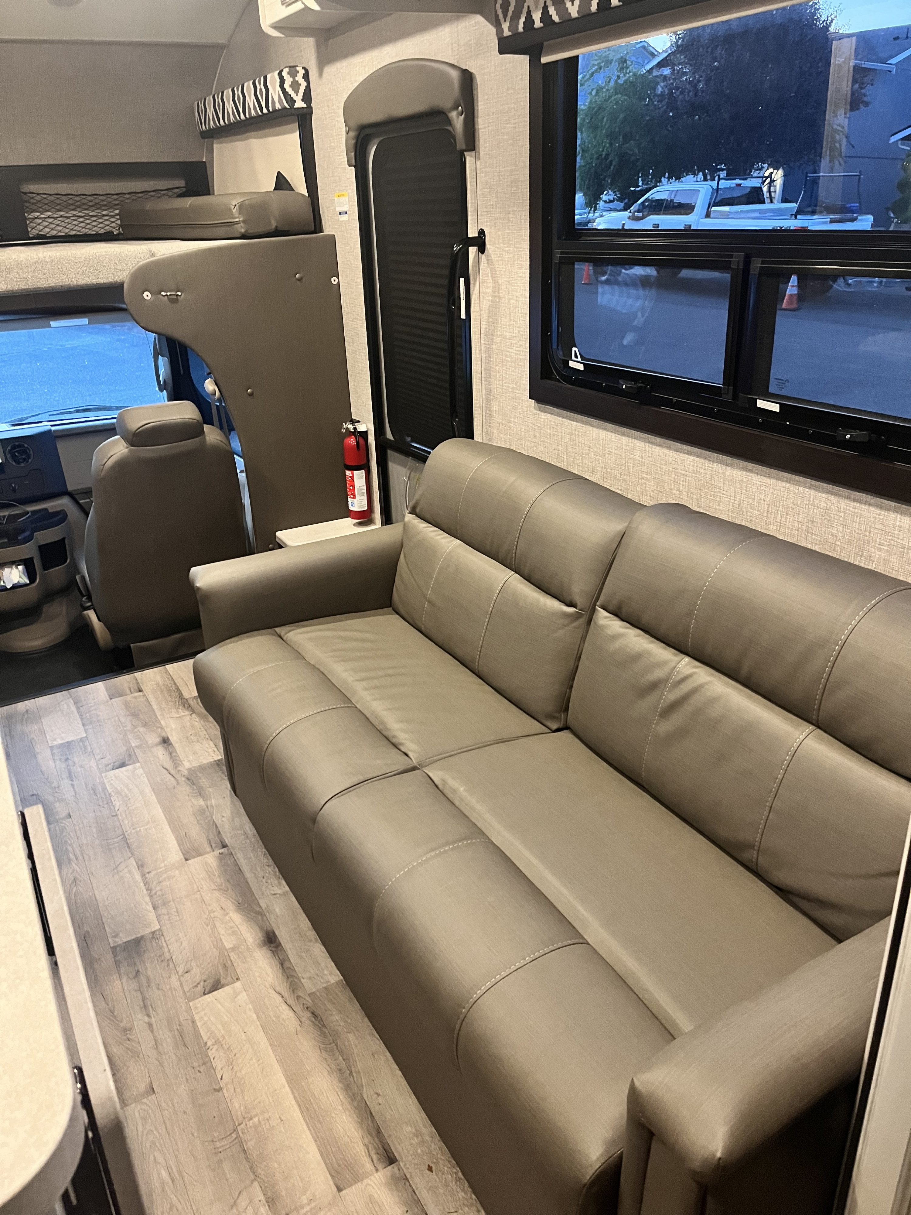 Spacious RV rental interior featuring a long sofa, light wood-look flooring, an overhead bunk, and driver's area.. Jayco Greyhawk 2022