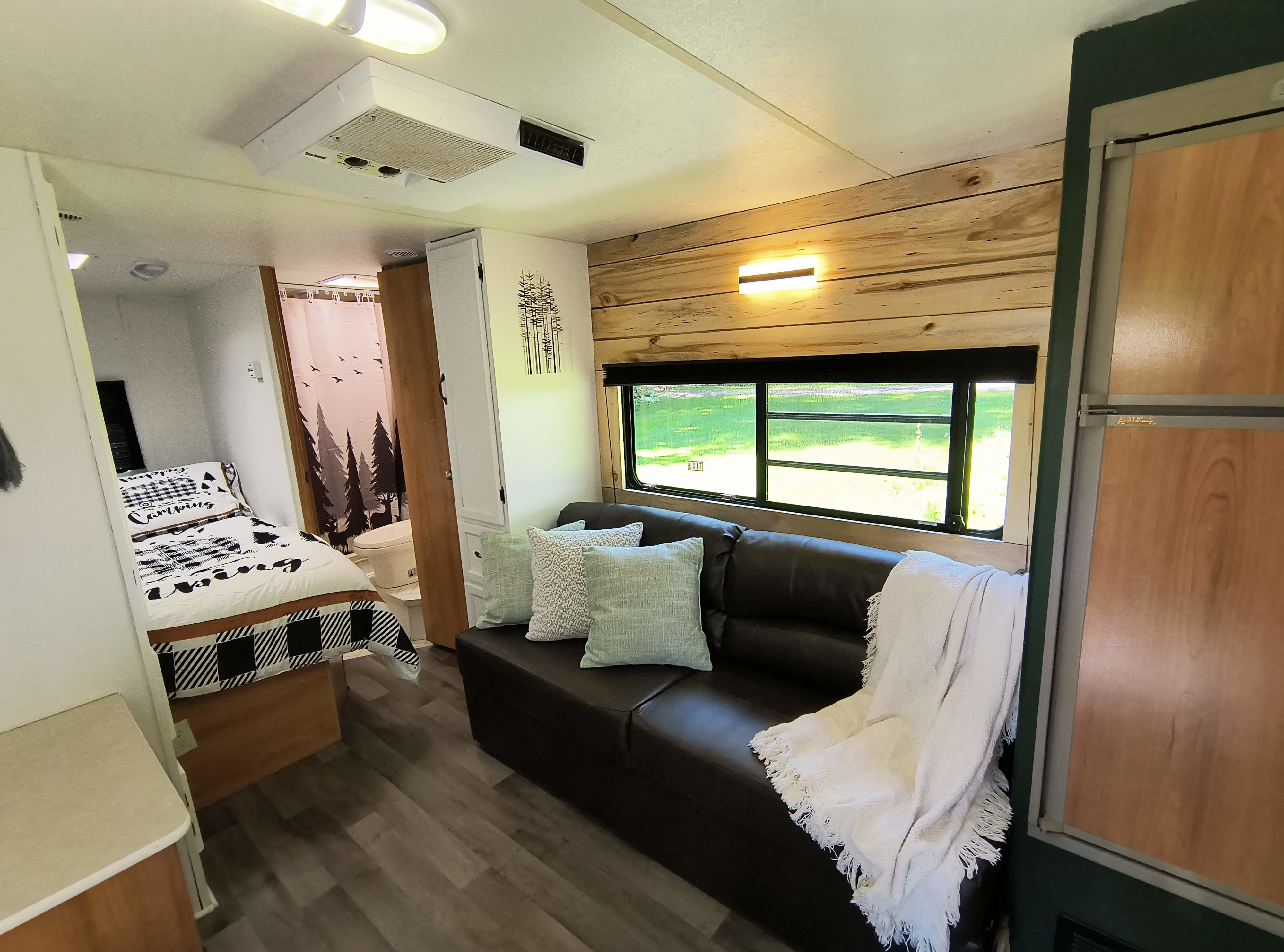 Cozy rv rental interior featuring a bed with buffalo plaid, dark sofa, rustic wood accent wall, and bright window view.. Coachmen Catalina 1998