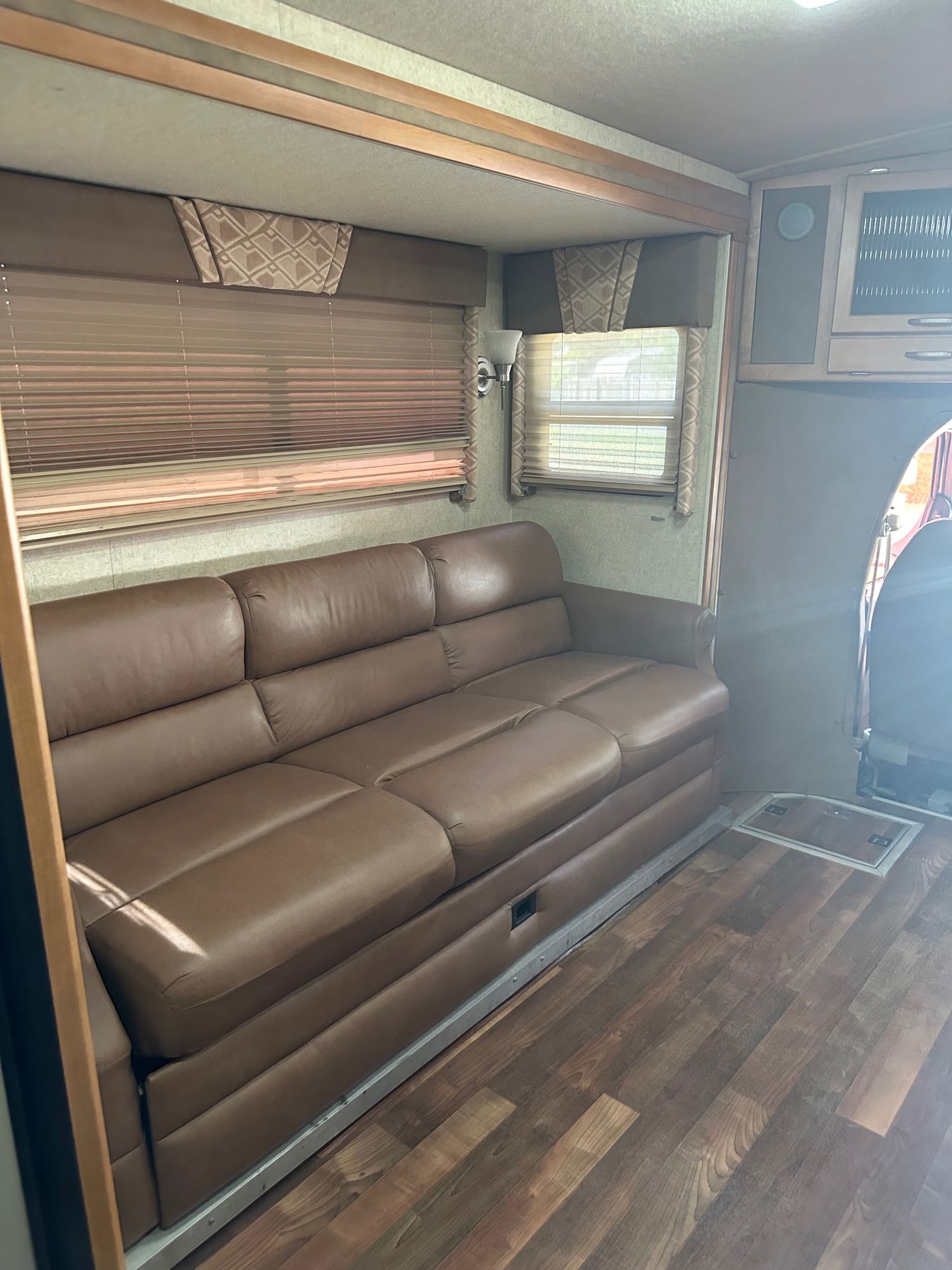 Spacious RV rental living area featuring a long brown leather sofa, wood-look flooring, and windows with blinds.. Winnebago Cambria 2008