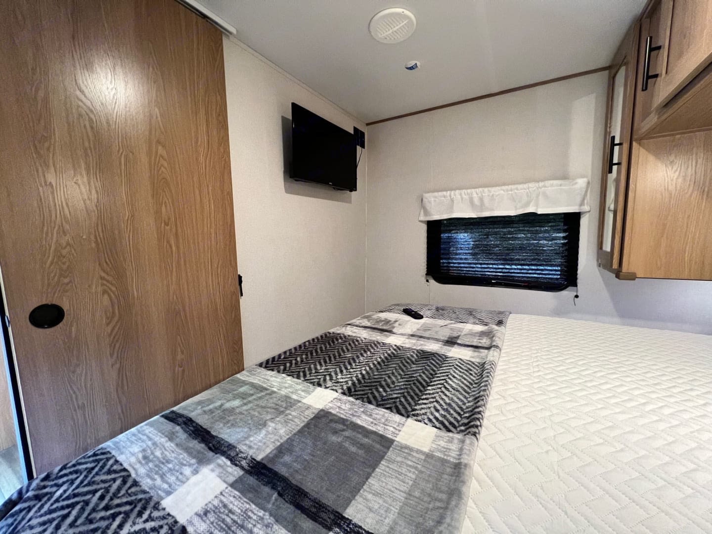 Interior of an RV rental bedroom showing a bed with plaid blanket, wall-mounted TV, window with blind, and wood sliding door.. Prime time Avenger 26BK 2023