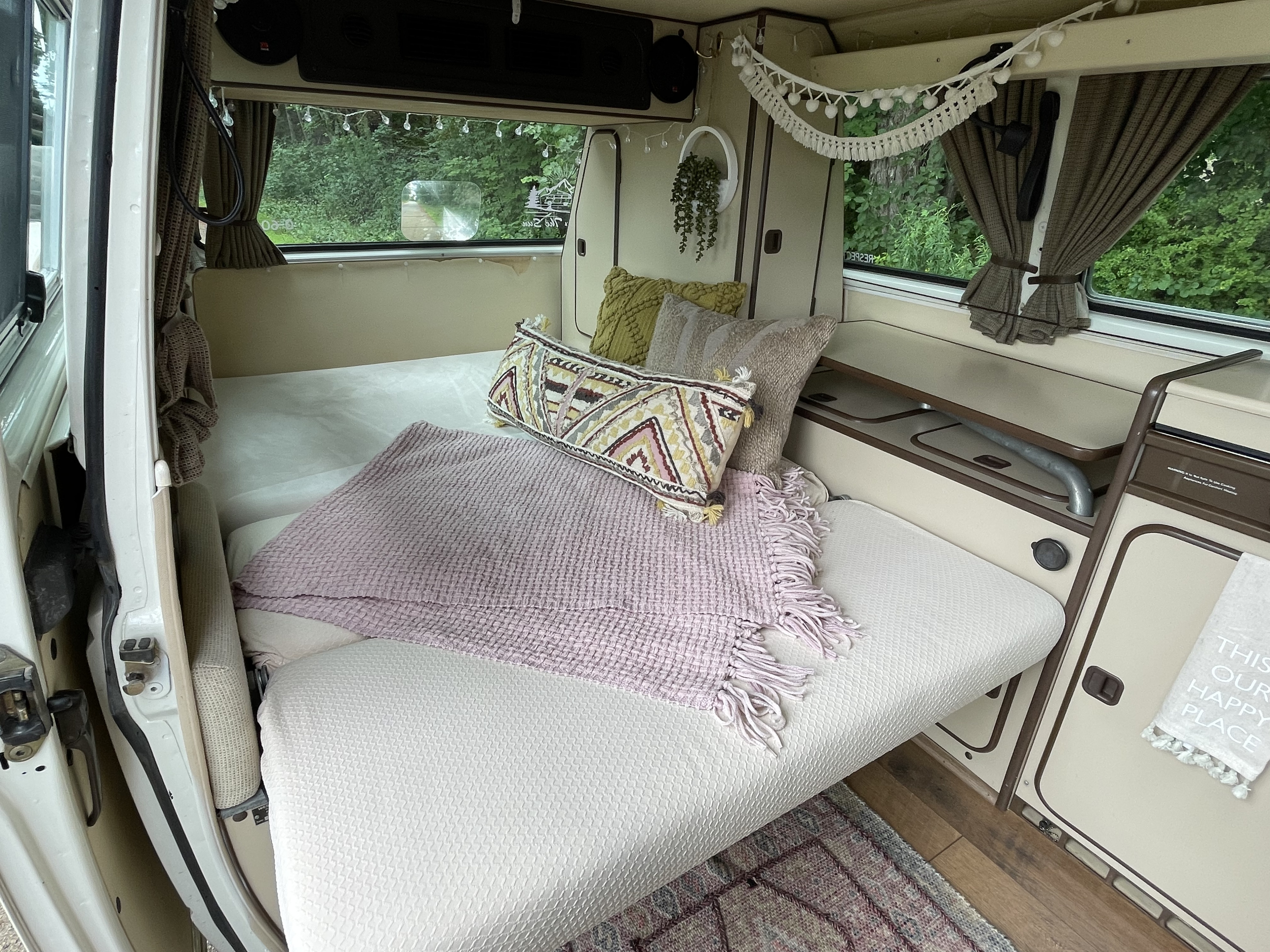 Cozy RV rental interior with a made bed, decorative pillows, and a pink blanket, overlooking green scenery.. Volkswagen Westfalia 1983