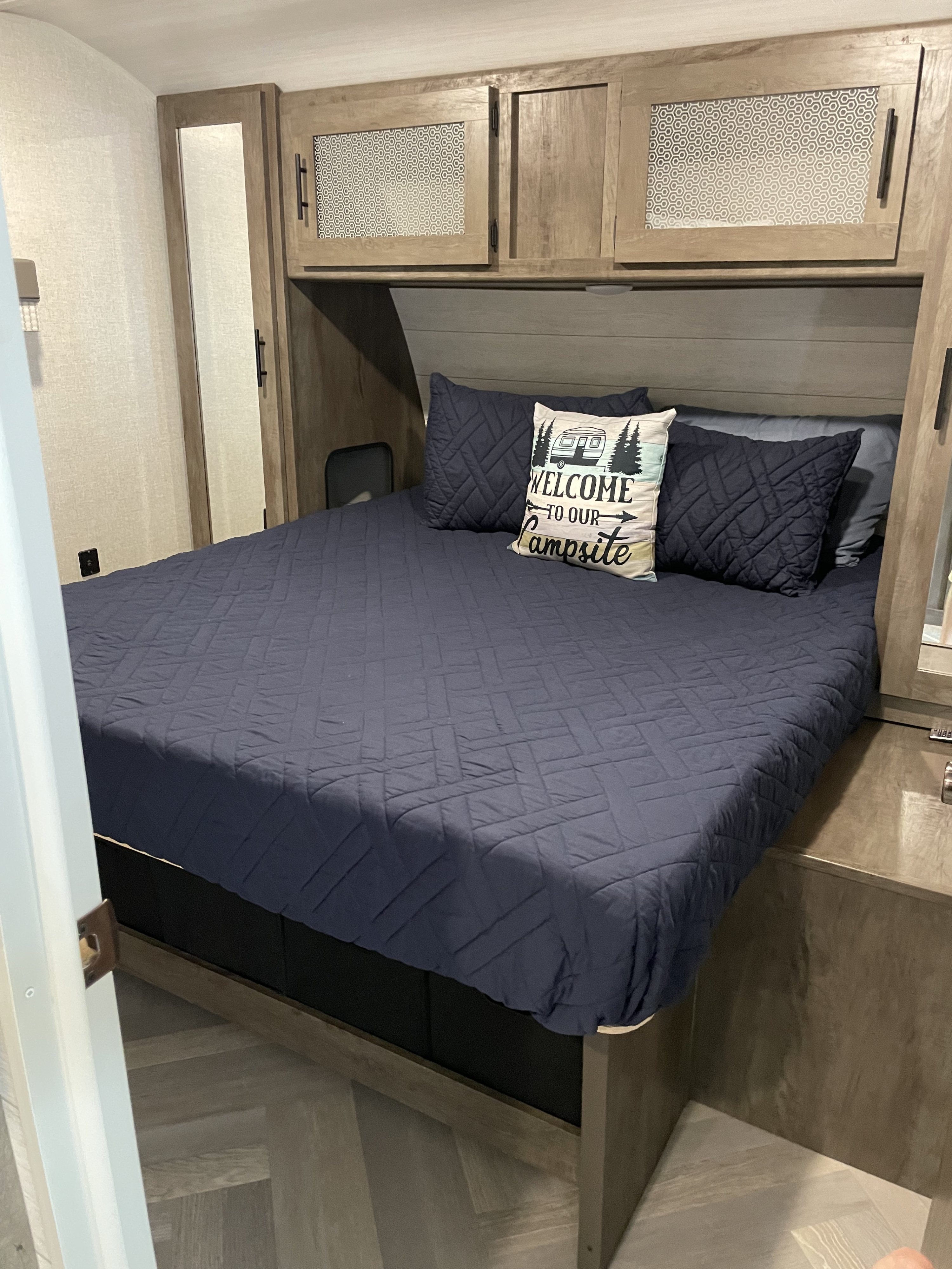 RV rental bedroom features a comfy bed with a "WELCOME TO OUR Campsite" pillow and ample overhead storage.. Forest River Wildwood 2021