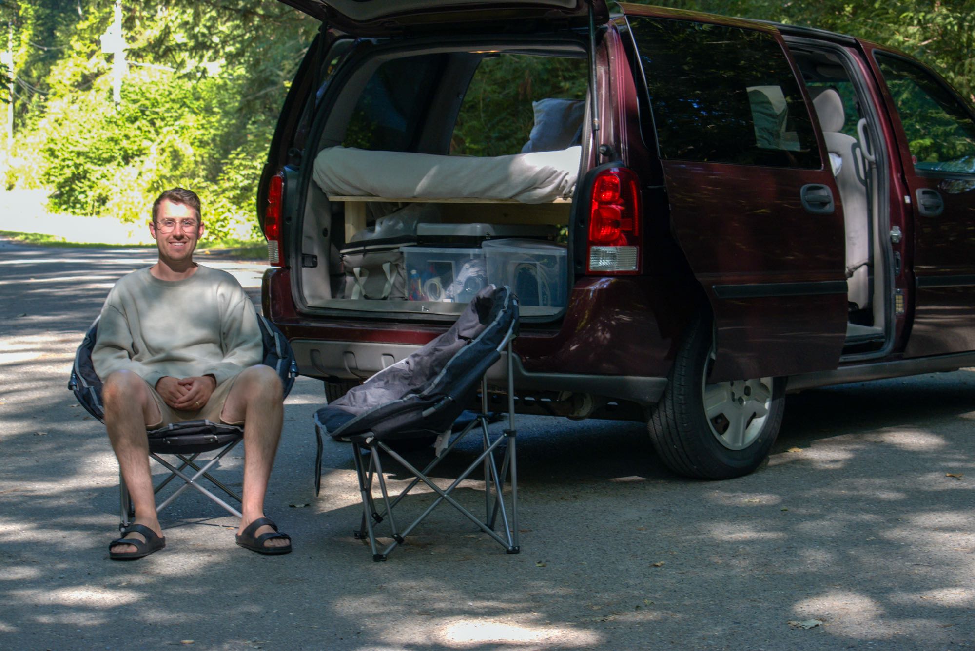 A man smiles, seated by his minivan, converted into an RV rental-style camper with a bed and storage for outdoor adventures.. Chevrolet Uplander 2008