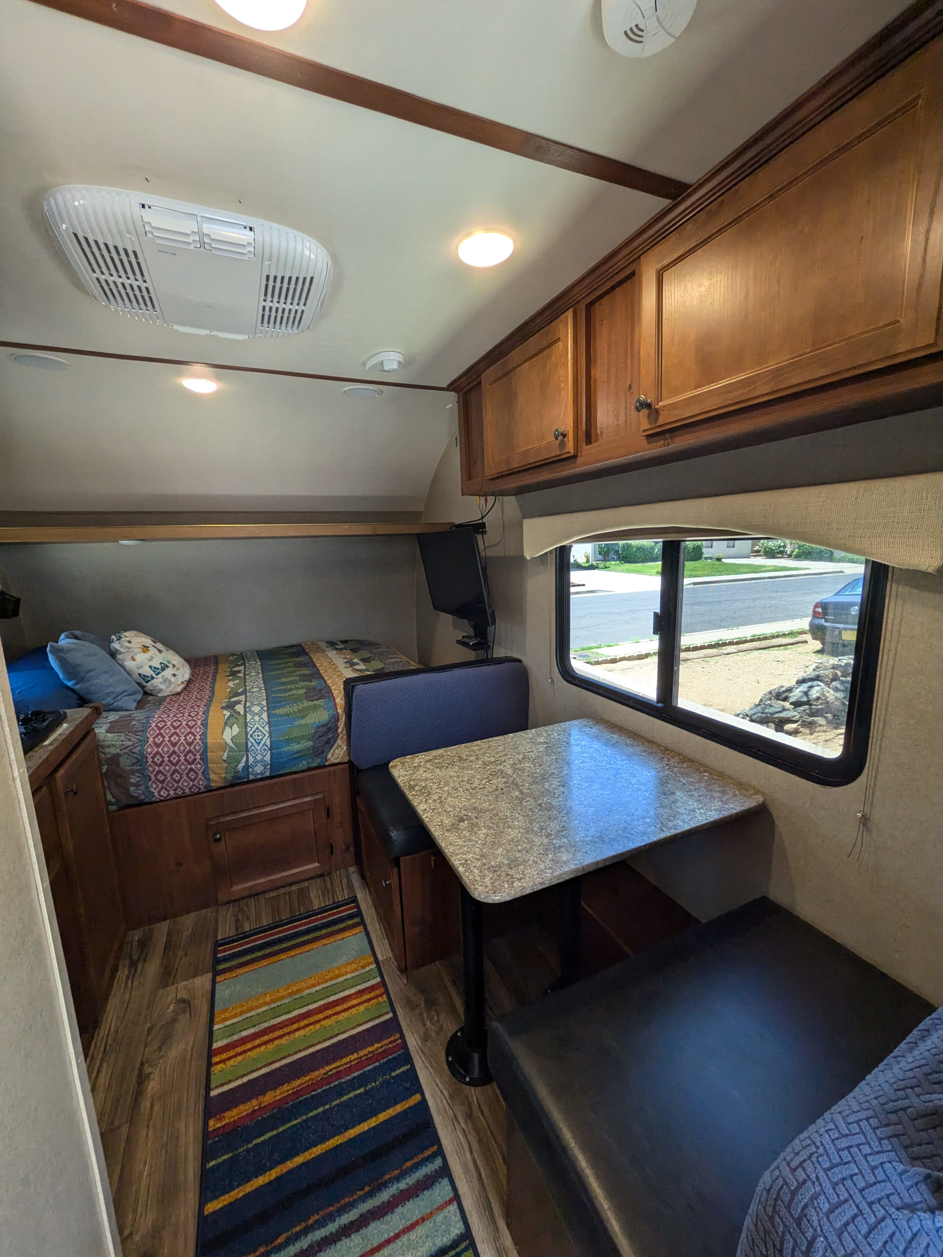 RV rental interior with a cozy bed, dinette, and wood cabinets. Features an AC unit, window view, and a colorful rug.. Sunset Park & Rv Inc. Sun-Lite 2019