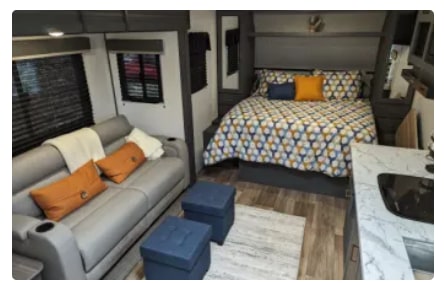 Cozy RV rental interior with a patterned bed, grey sofa & orange pillows, plus a compact kitchen. Ideal for mobile living.. Keystone RV Bullet Crossfire 2023