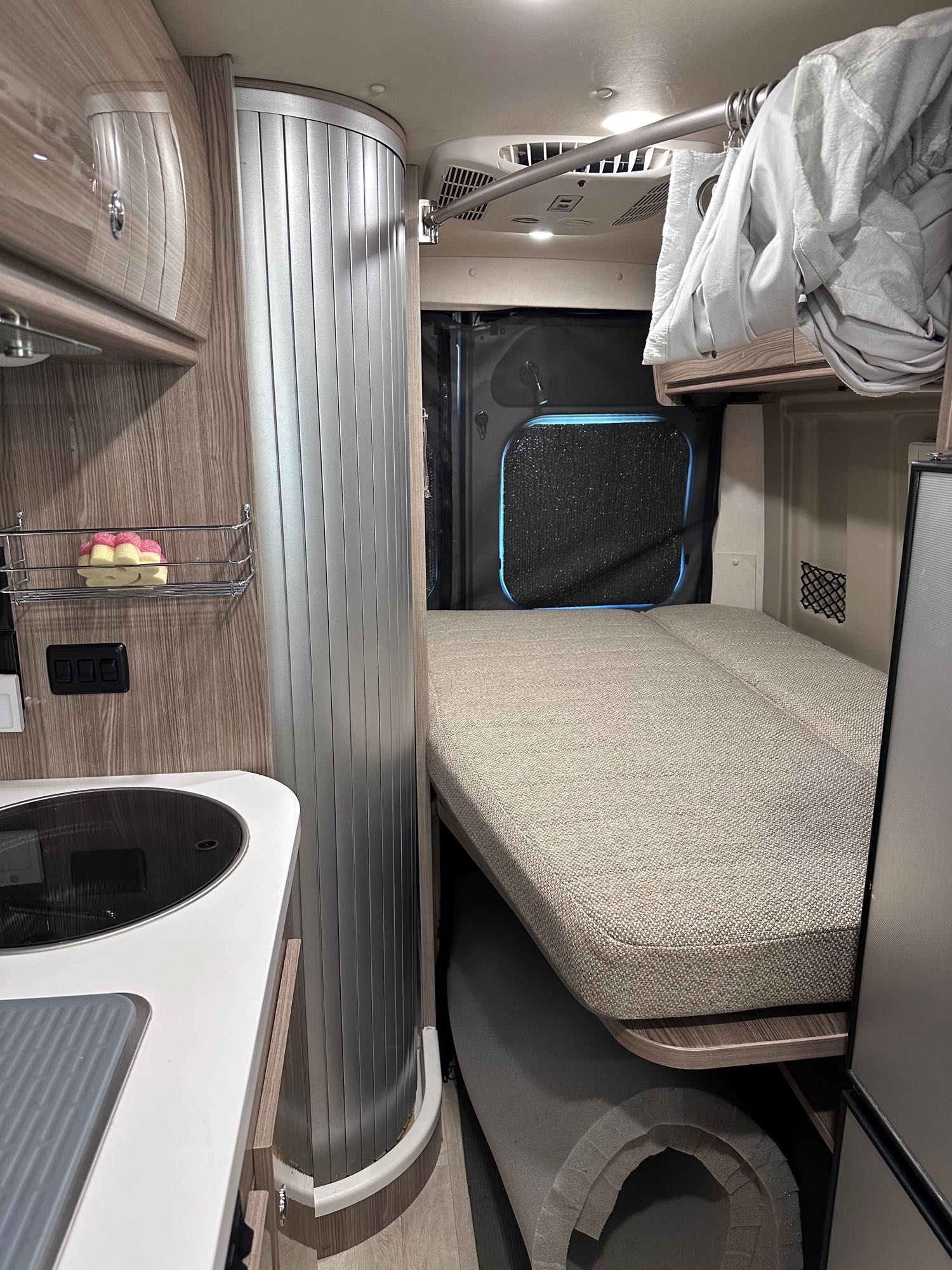 RV rental interior: A compact living space with a kitchen counter on the left, a curved door, and a comfy bed at the back.. Winnebago Travato 2022