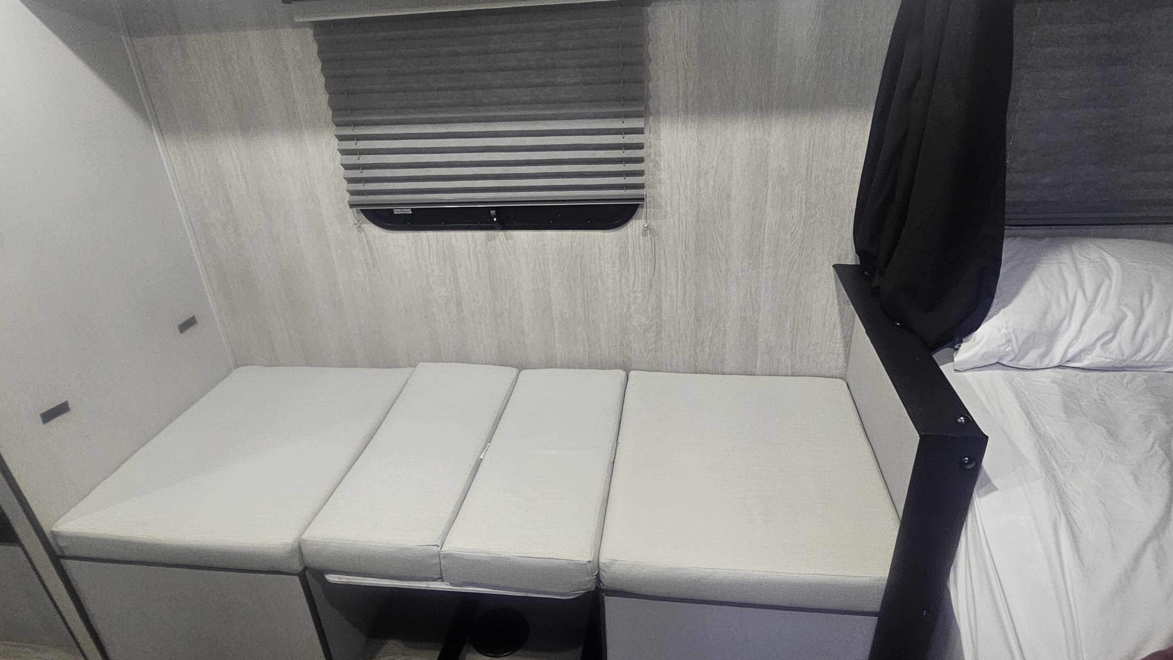 RV rental interior: convertible seating/bed with light grey cushions, a window with blinds, and an adjacent bed.. Genesis Supreme Seabreeze Toy Hauler 2024
