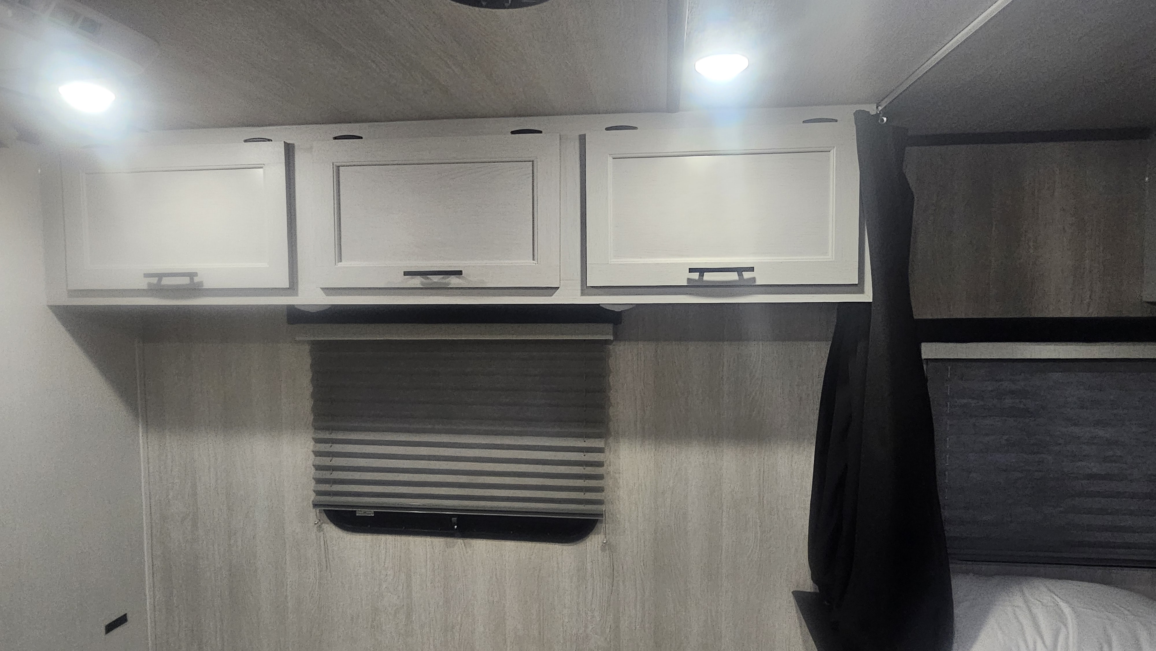 An RV rental bedroom with practical overhead storage, bright lighting, and a window with privacy blinds.. Genesis Supreme Seabreeze Toy Hauler 2024