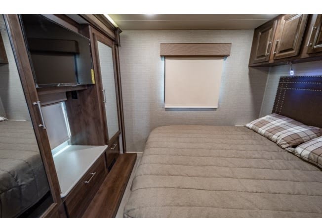 Inside an RV rental, a bedroom with a quilted bed, plaid pillows, built-in TV, dark wood storage, and a window.. Entegra Coach Vision XL 2023