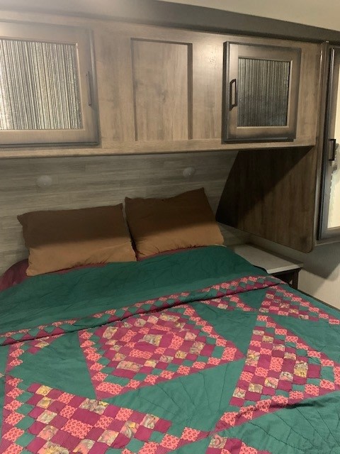 Inside this RV rental's cozy bedroom: a green & pink patterned quilt, brown pillows, and overhead storage cabinets.. Heartland RVs Mallard 2021