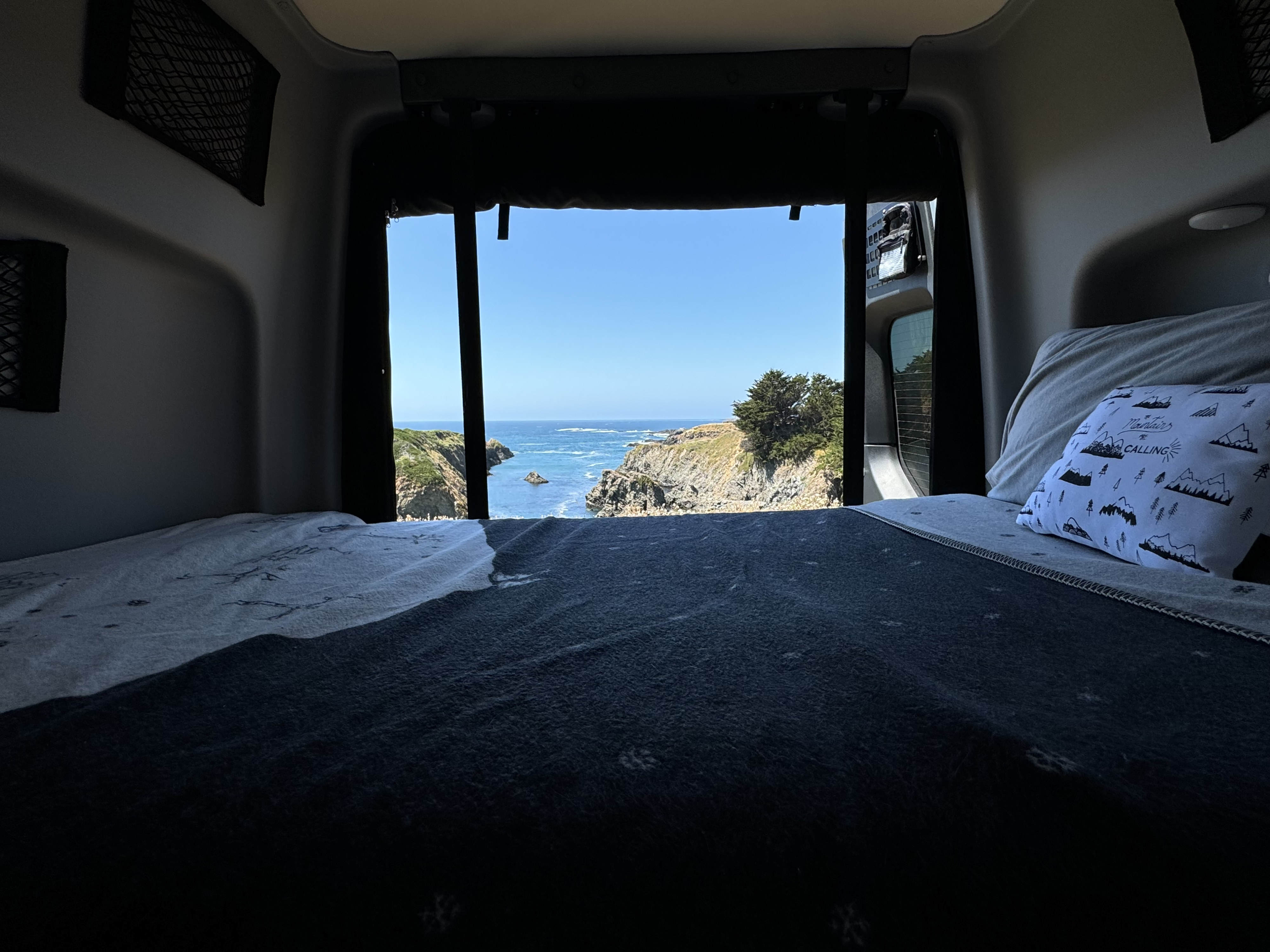 Inside an RV rental: a bed offers stunning ocean views of cliffs & blue water from open back doors.. Winnebago Revel 2022