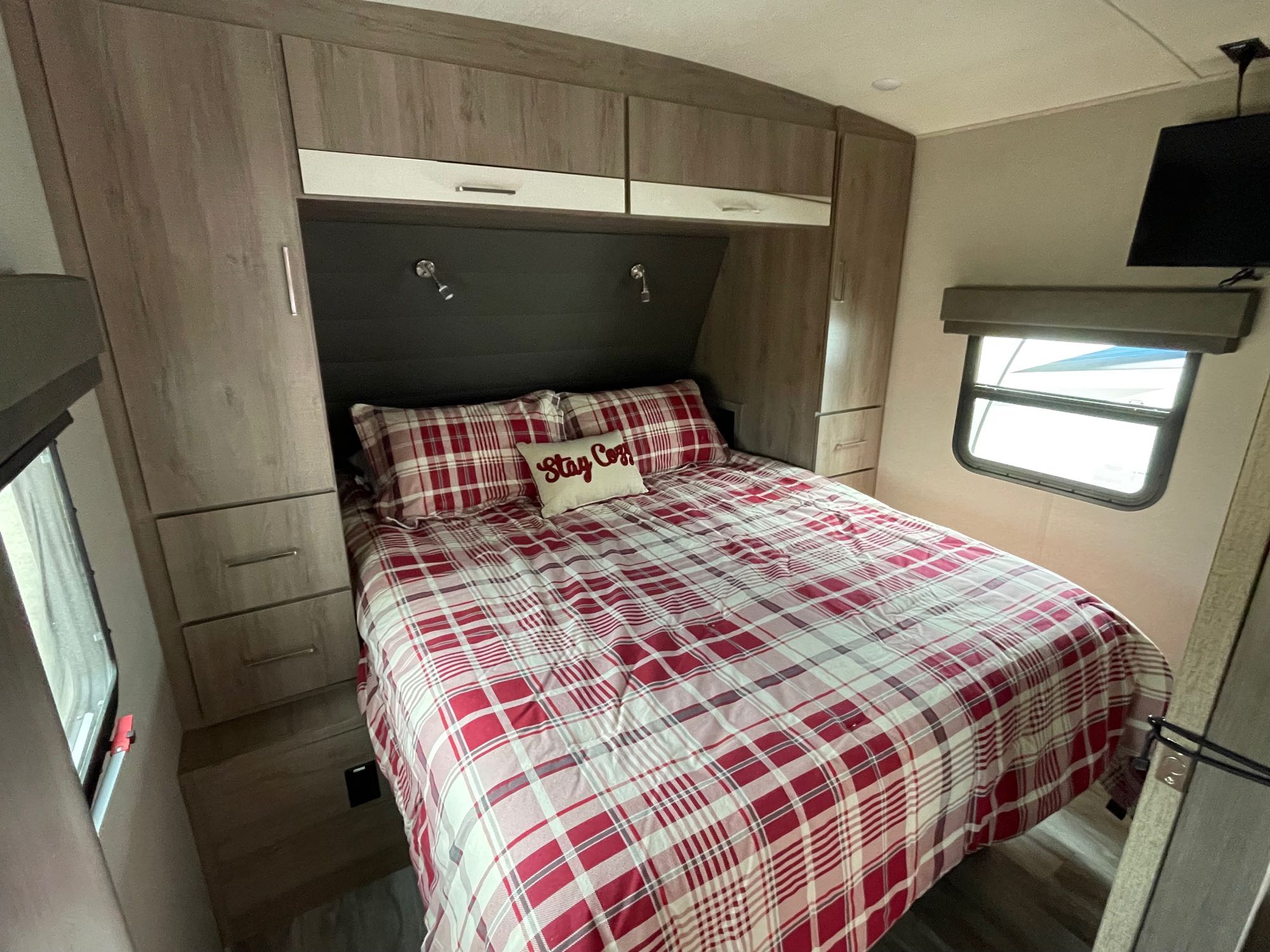 This RV rental bedroom features a plaid bed, ample built-in storage, a TV, and a window, offering a cozy retreat.. Grand Design Imagine 2022