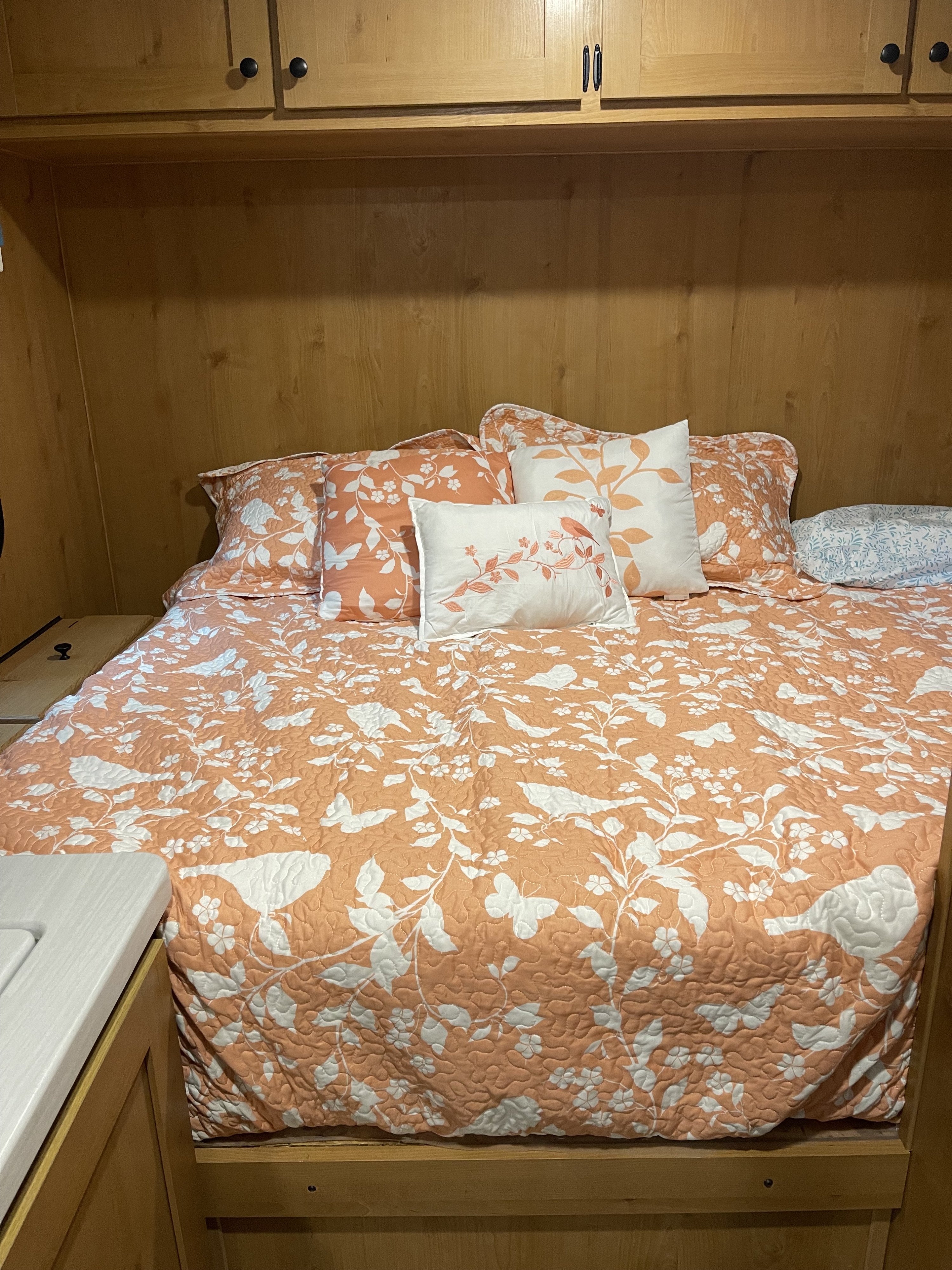 RV rental interior: cozy bedroom featuring a peach-patterned quilt, decorative pillows, and overhead wooden cabinets.. Riverside Retro Elite 2024