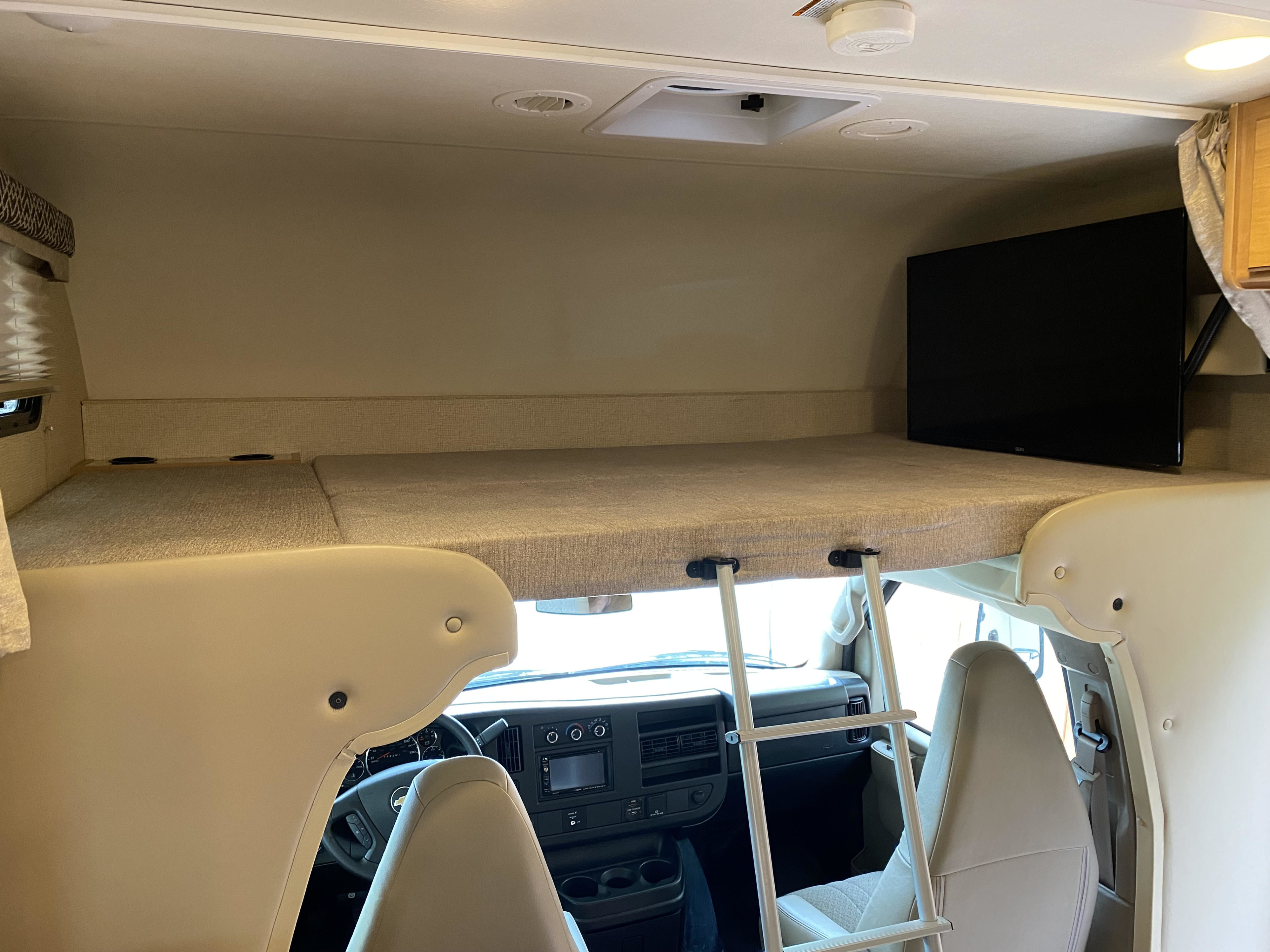 Inside an RV rental: a comfortable cab-over bunk bed with a TV, ladder access, and front cockpit view.. Thor Four Winds 2017