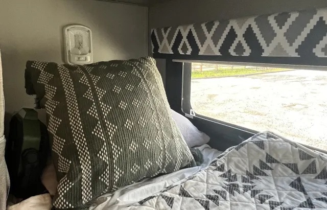 Cozy RV rental bed with a green patterned pillow, geometric quilt, and window view with a valance.. Jayco Greyhawk 2021