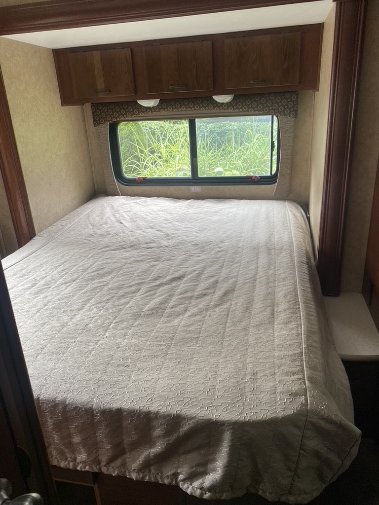Cozy RV rental bedroom featuring a large bed with patterned comforter, window viewing nature, and overhead wood cabinets.. Forest River Sunseeker 2014