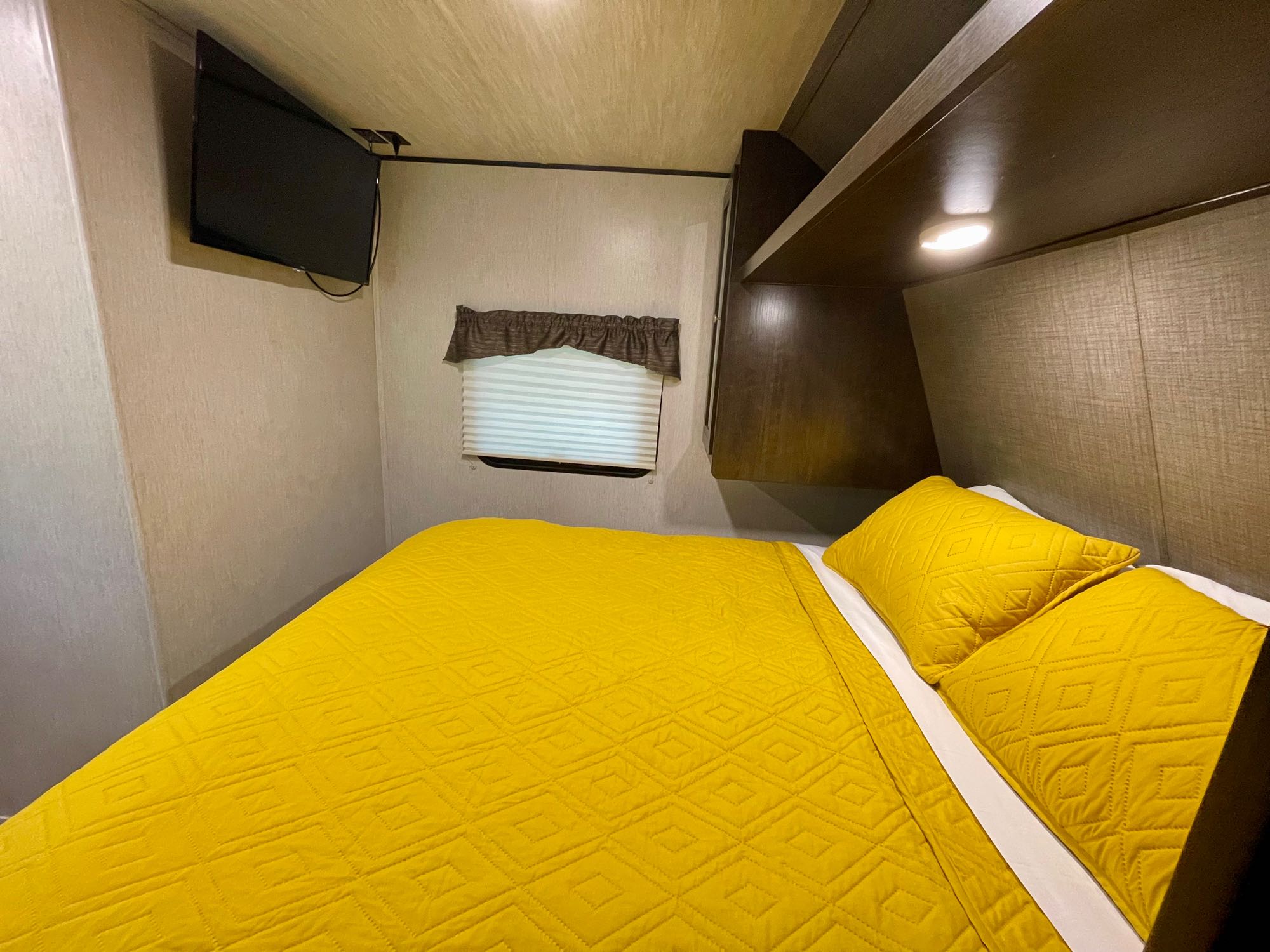 Bright and cozy RV rental bedroom. Features a yellow quilted bed, mounted TV, window with blind, & overhead lighting/storage.. Heartland RVs Trail Runner SLE 2019