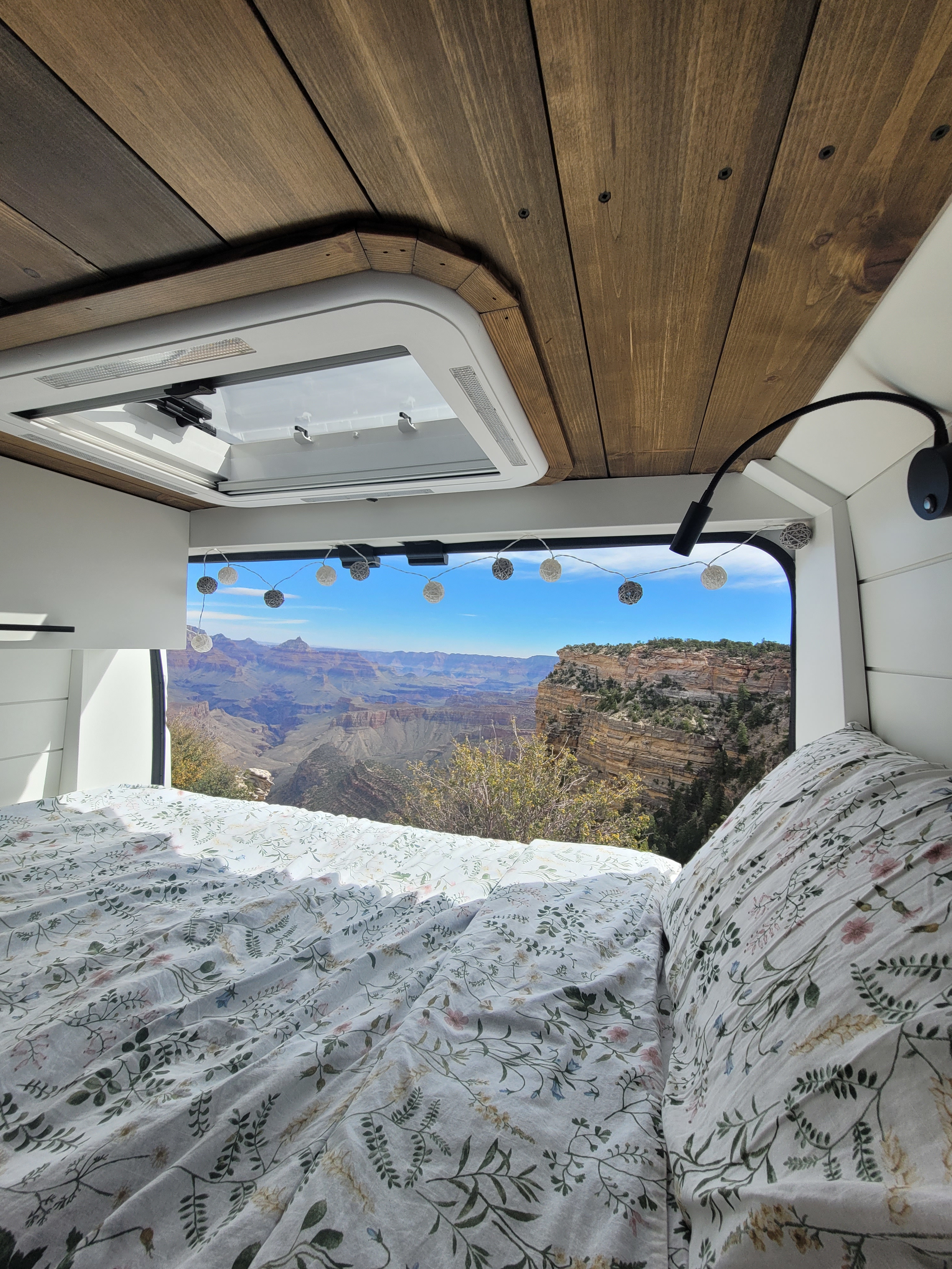 Cozy RV rental bed with floral sheets overlooking the Grand Canyon. Wood ceiling & string lights add charm.. Ram Promaster 3500 159 Ext 2022