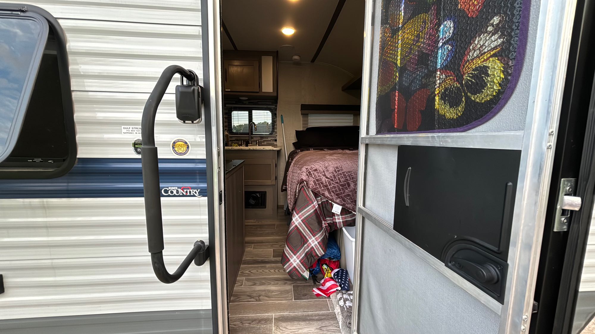 Open RV rental door reveals cozy interior: bed, kitchen, and patterned blankets. Ideal for camper travel.. Gulf Stream Ameri-Lite Super Lite 2021