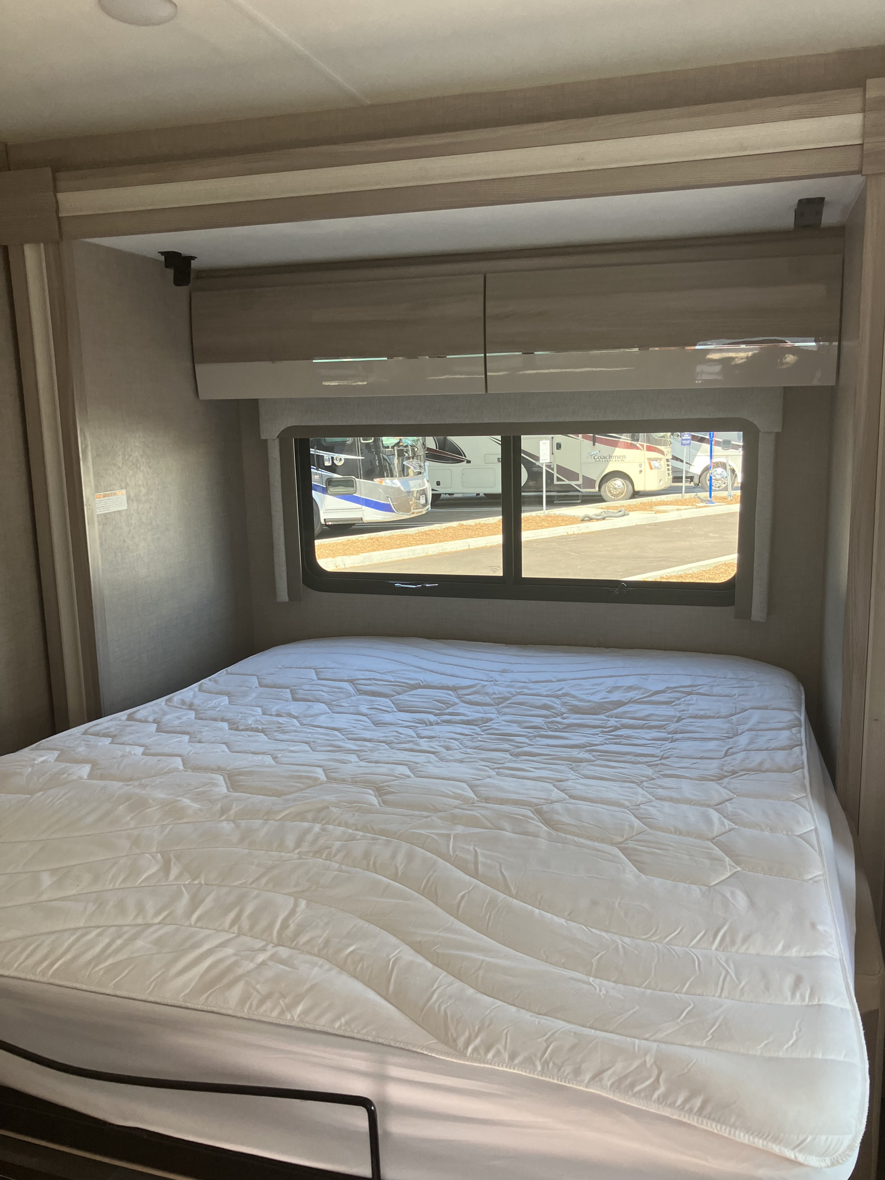 Inside this RV rental: a bedroom with a white bed, overhead cabinets, and window showcasing other RVs outside.. Thor Motor Coach Compass 2022
