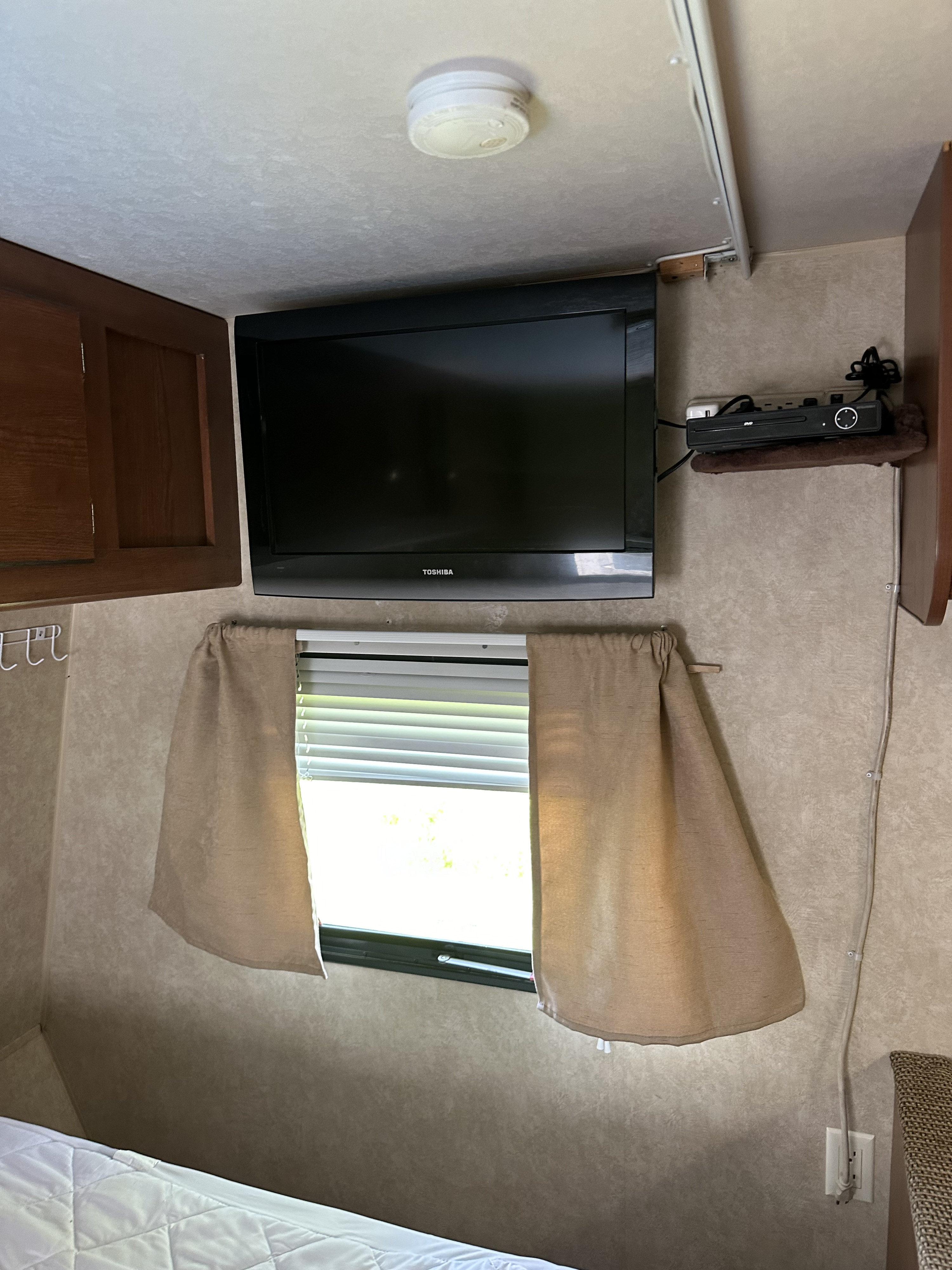 Compact RV rental bedroom with wall-mounted TV, DVD player, and window with blinds and curtains.. Forest River Salem Cruise Lite 2014