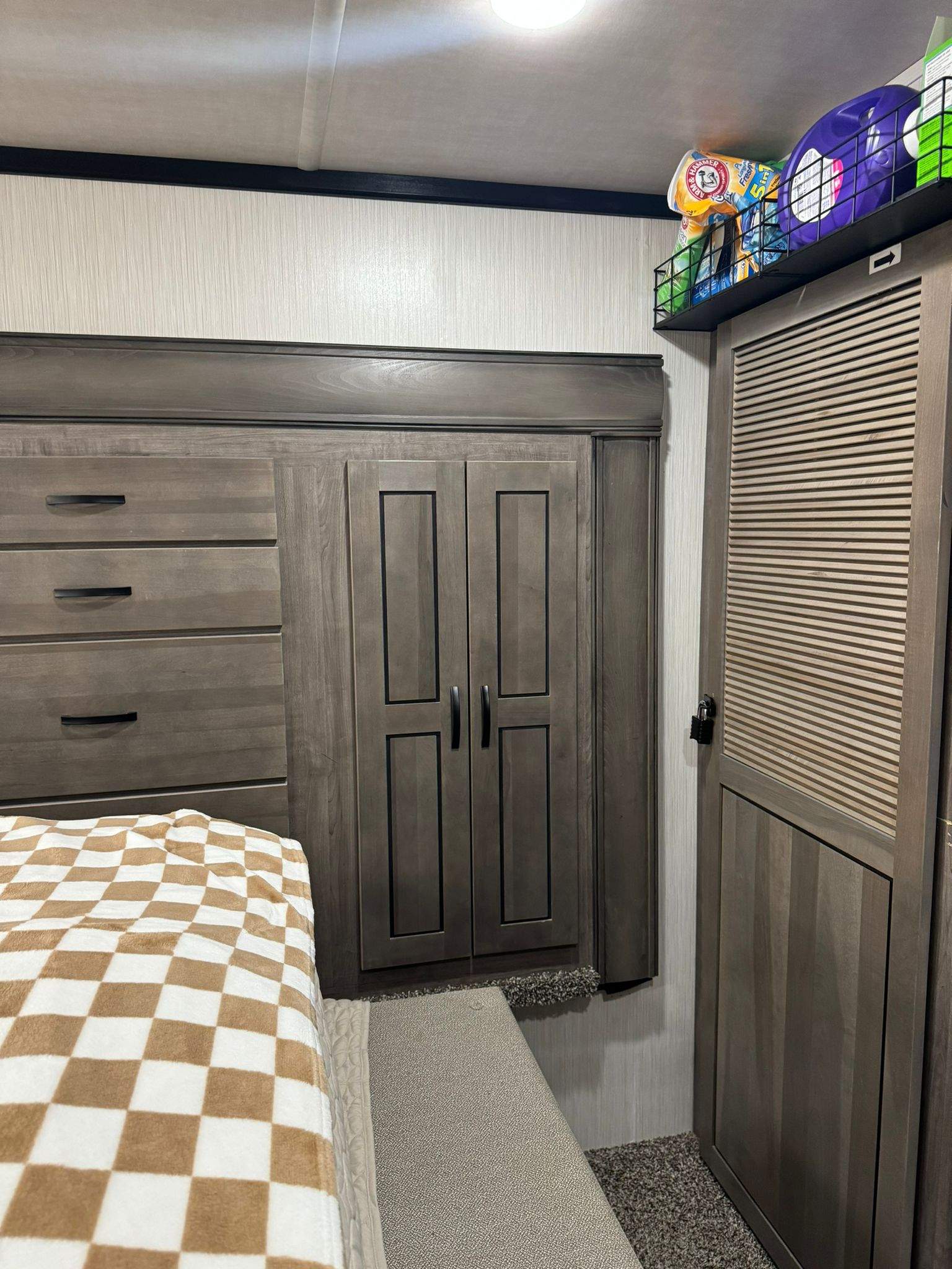 Cozy RV rental bedroom: bed with checkered blanket, dark wood dresser, wardrobe, and a shelf with laundry supplies.. Cedar Creek Cedar Creek Fifth Wheel 2022