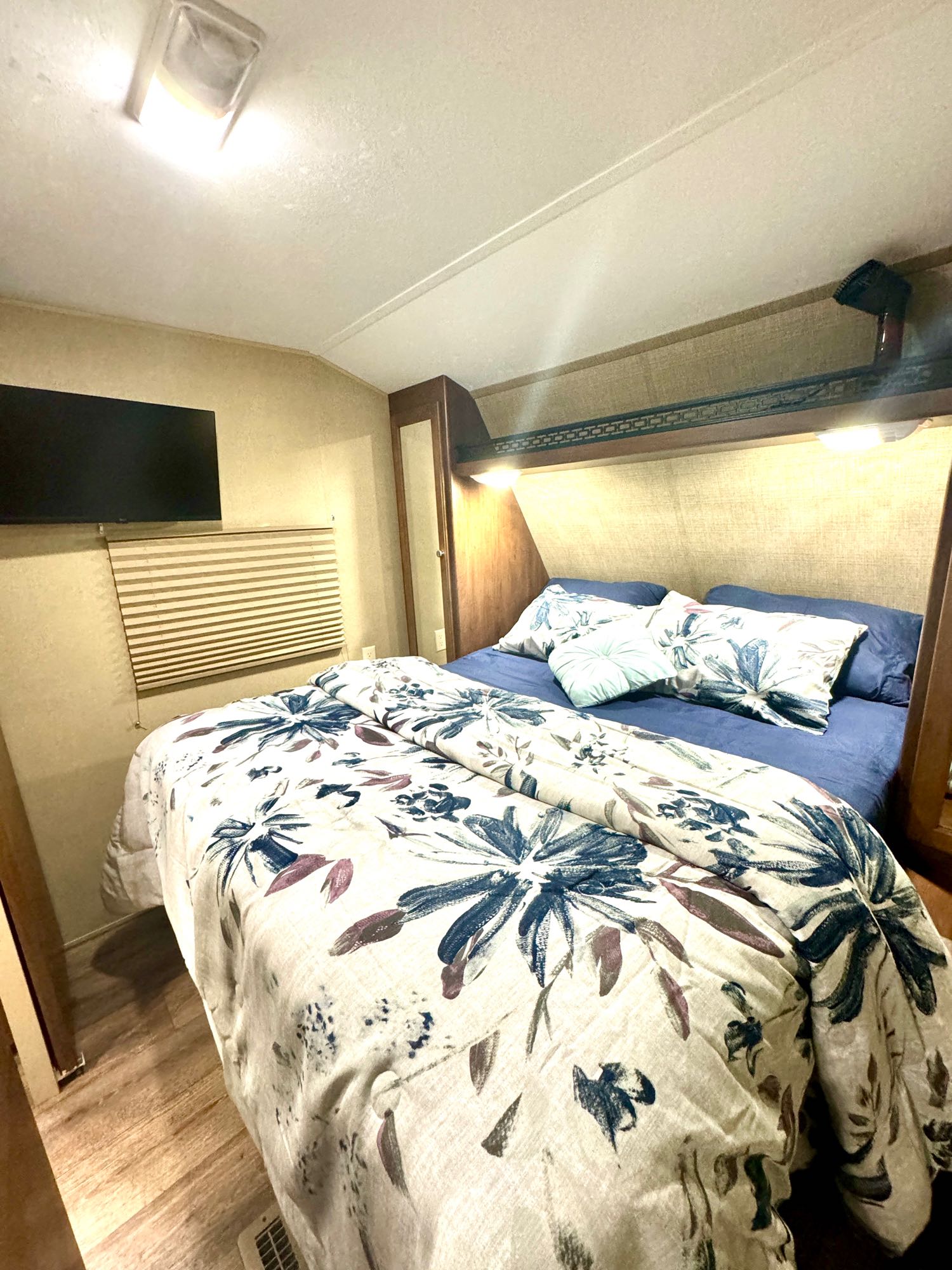 Inside an RV rental: a compact bedroom featuring floral bedding, wall-mounted TV, and bright overhead lights.. Aspen Trail Aspen Trail Trailer 2014