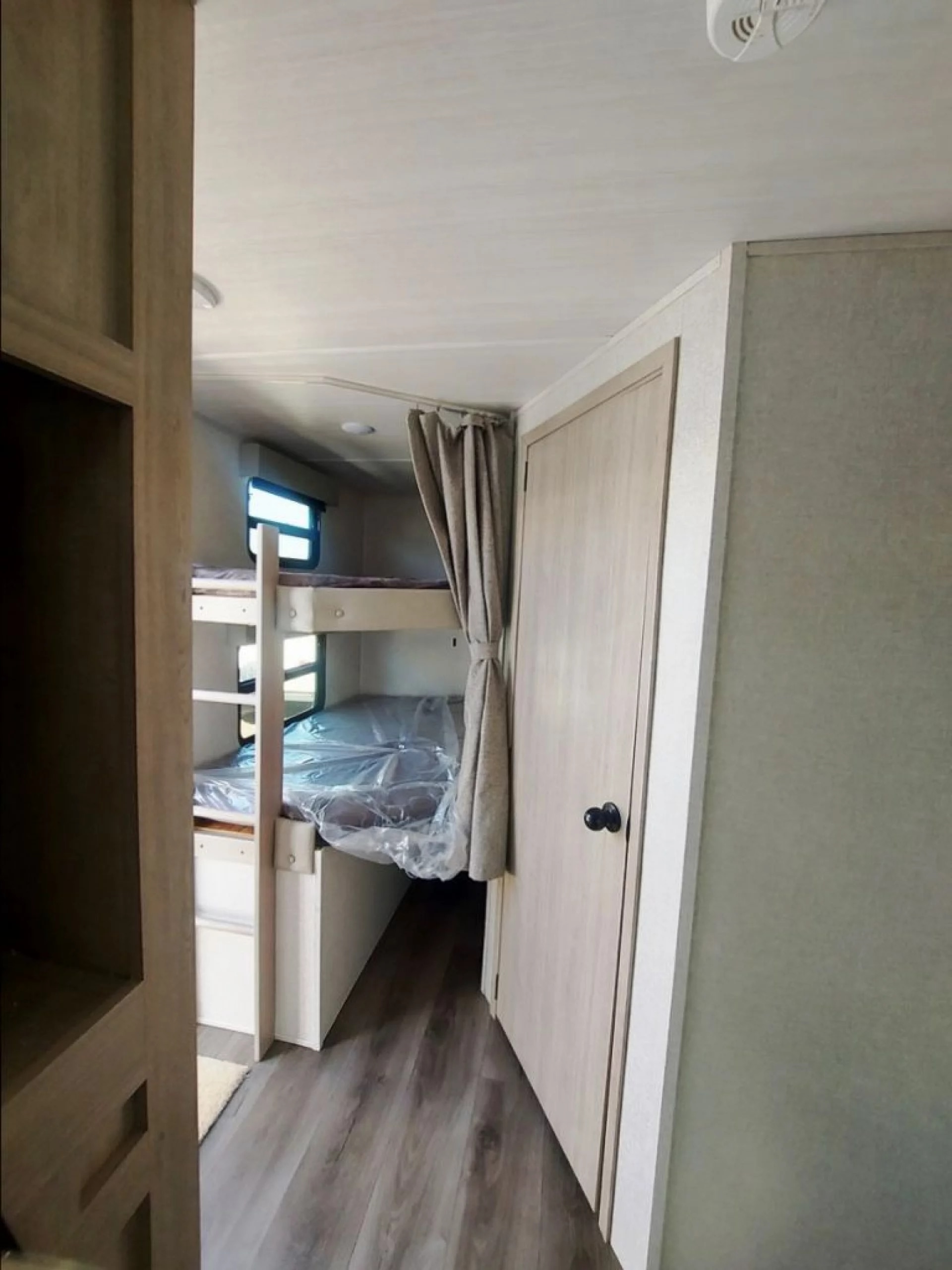 RV rental with clean bunk beds on the left, still covered in plastic. A privacy curtain is drawn, next to a light-colored door.. Forest River East to West 2022