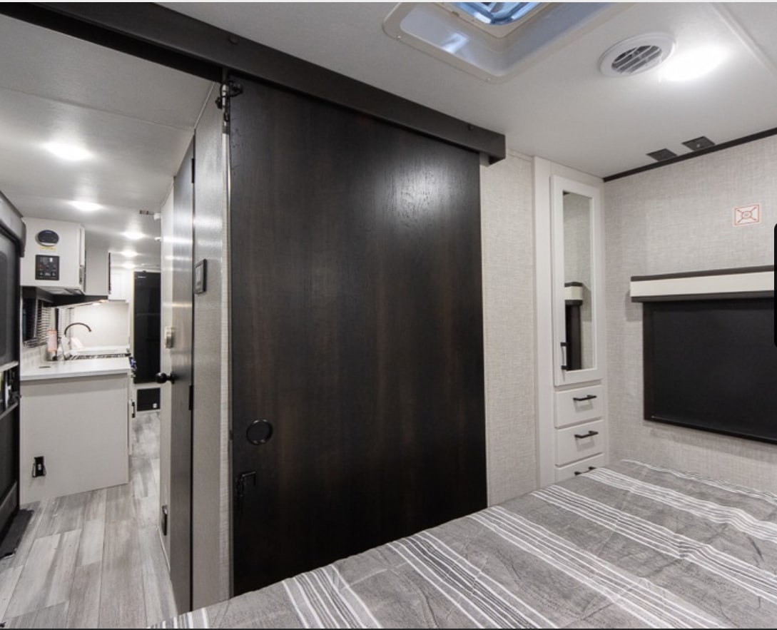 RV rental interior showing a bedroom with a grey striped bed, white cabinet, and a dark sliding door opening to a kitchen area.. Jayco Jay Feather 2024