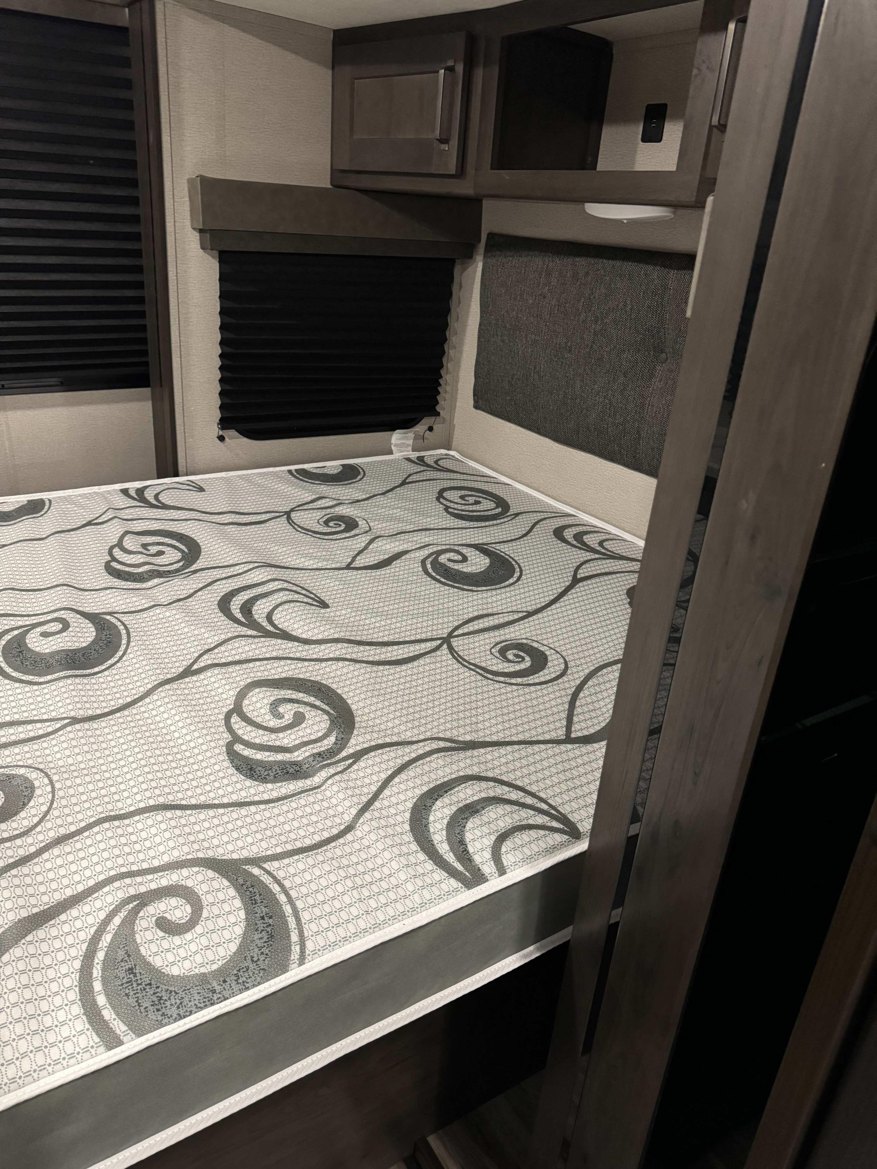 RV rental bedroom with patterned mattress, pleated window blinds, and overhead cabinets. A ladder is to the right.. Grand Design Transcend Xplor 2023