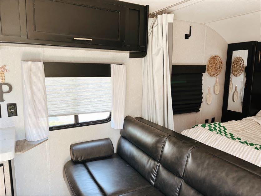 RV rental living space: dark cabinets, window with pleated blinds, and a brown sofa, next to a curtained bedroom.. Forest River Cherokee Grey Wolf 2021