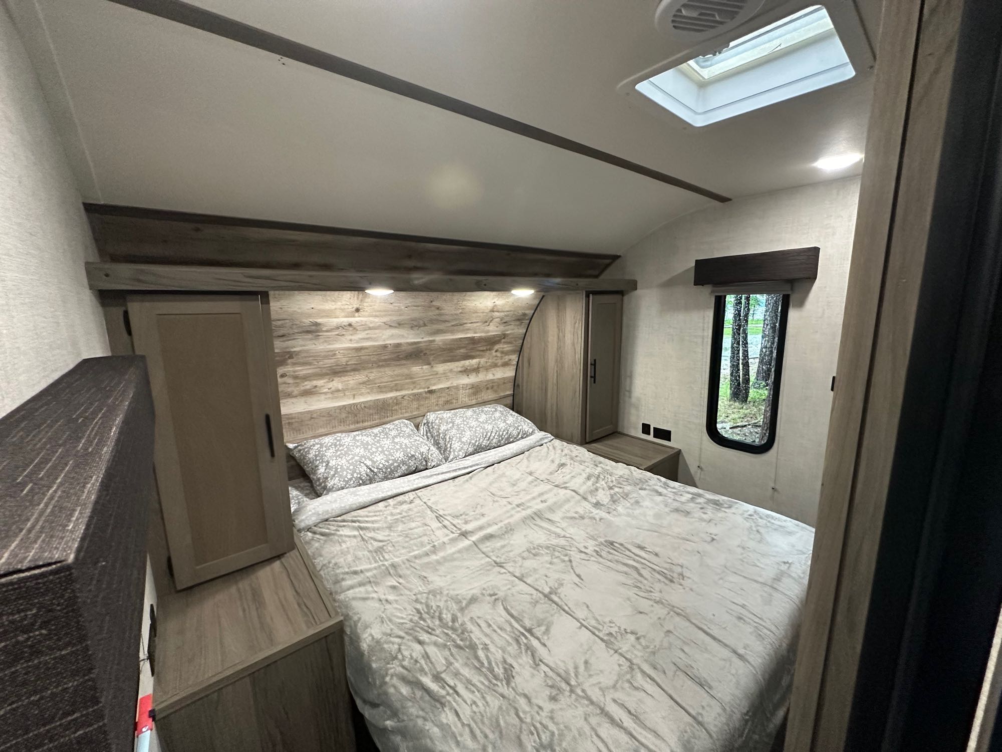 Inside a cozy RV rental bedroom: queen bed, storage, and a forest view from the window.. Gulf Stream Ameri-Lite 2021