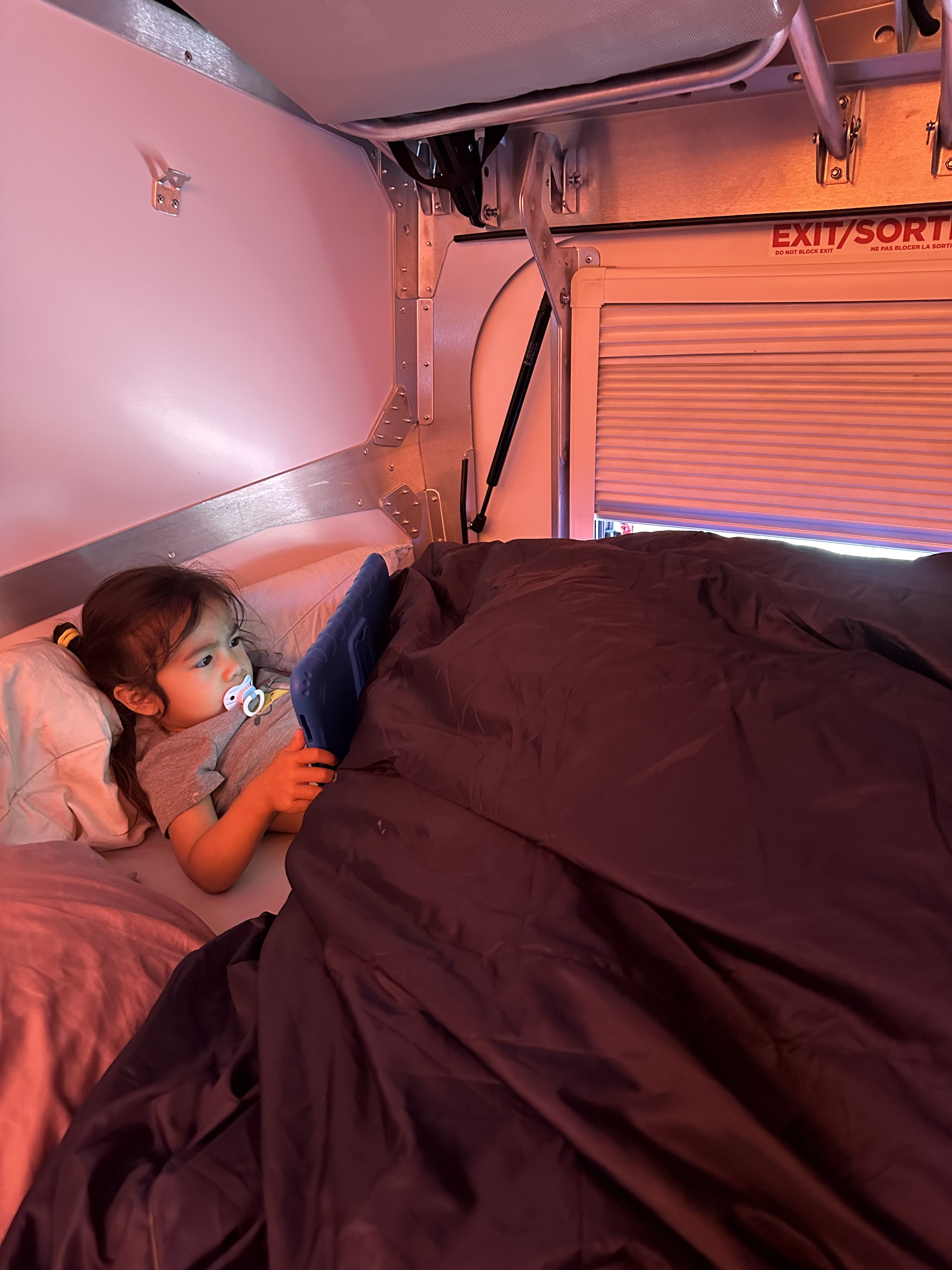 Inside an RV rental, a child in bed uses a tablet under warm light, with an "EXIT/SORTIE" sign nearby.. TAXA Outdoors Cricket Camper Overland Edition 2024