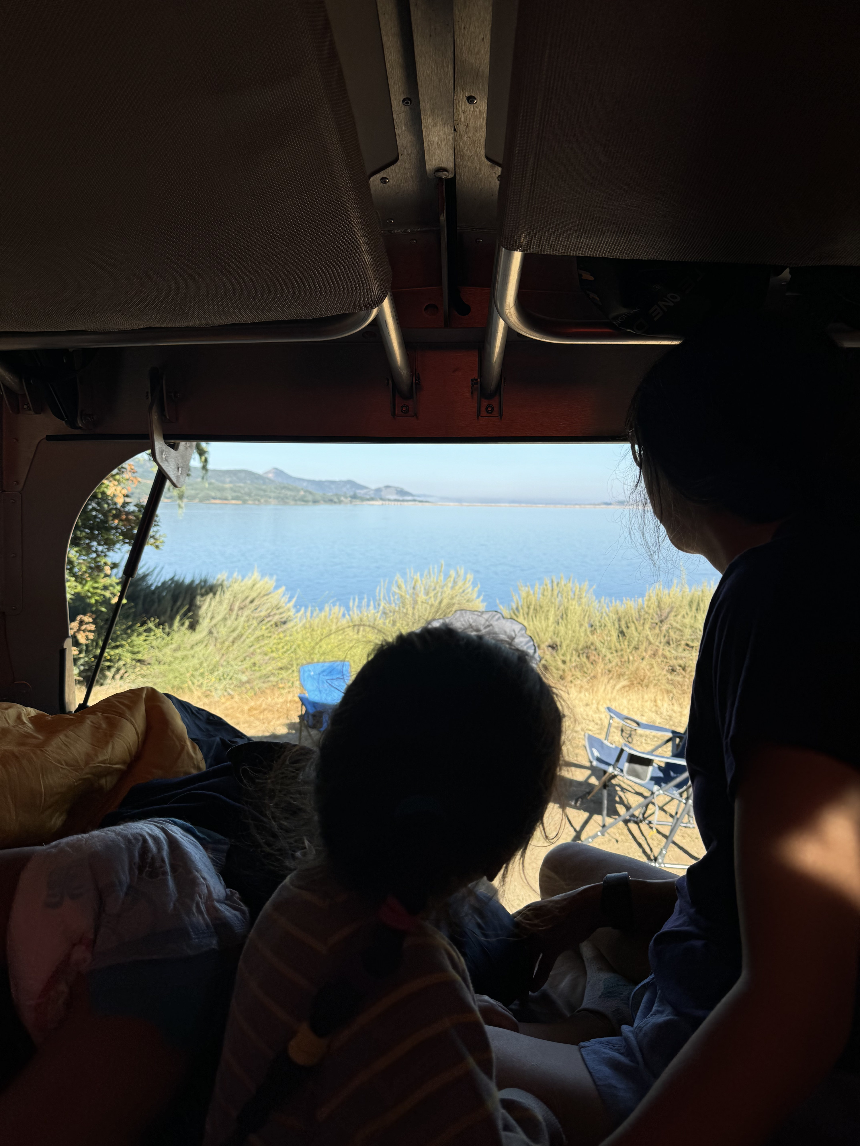 Inside our RV rental, an adult & child gaze at a serene lake & mountains. A peaceful camping view.. TAXA Outdoors Cricket Camper Overland Edition 2024