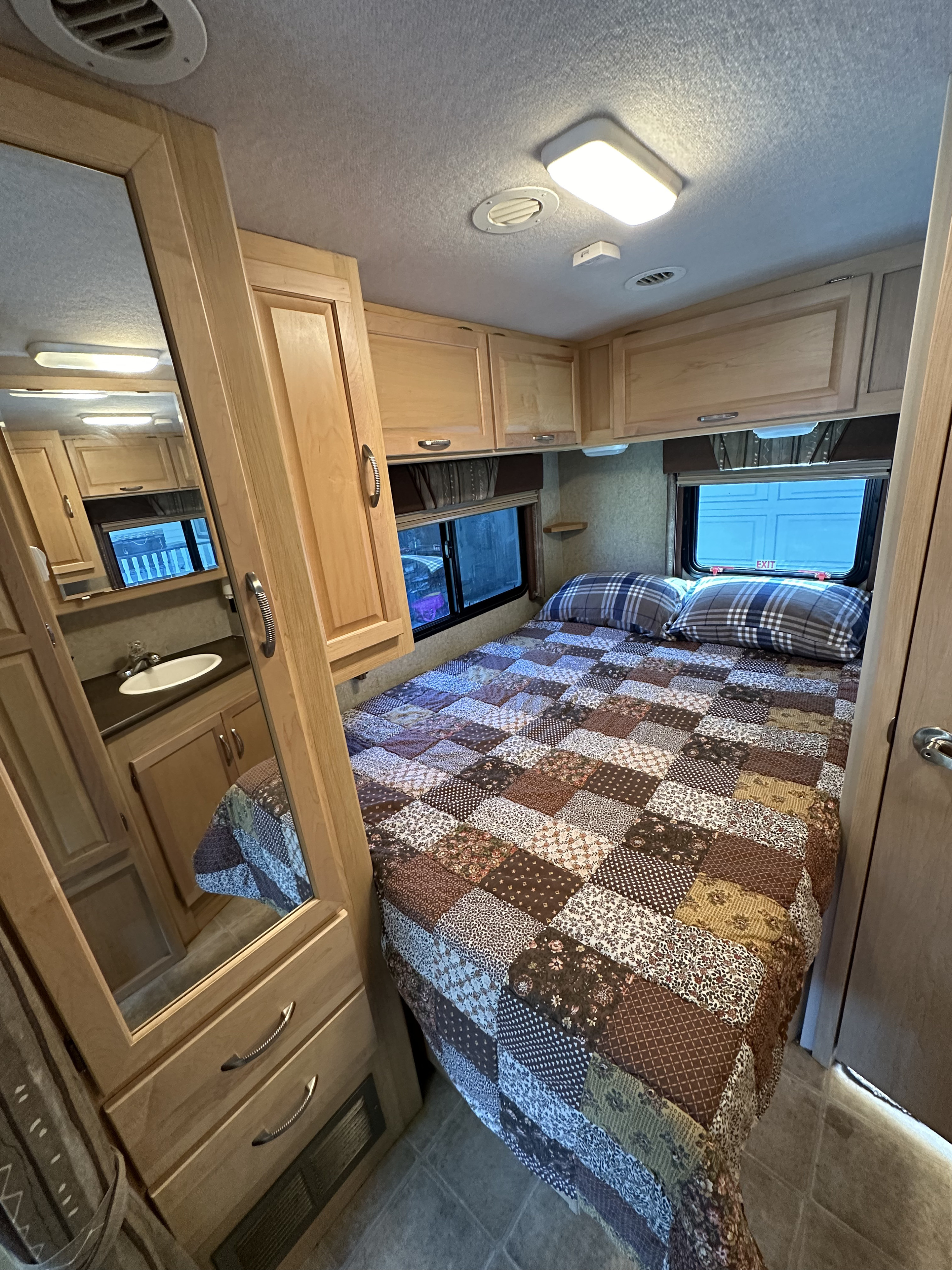 An inviting RV rental bedroom with a patchwork quilt, plaid pillows, overhead cabinets, and a mirror reflecting a bathroom sink.. Itasca Cambria 2008