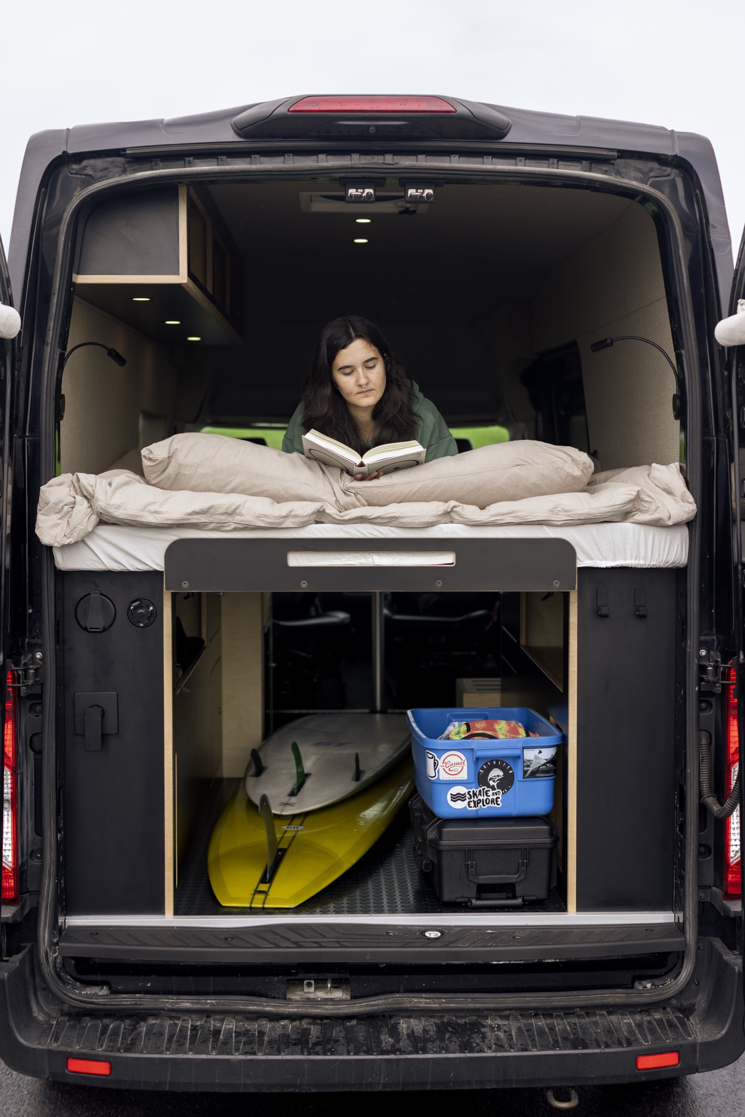 Enjoying an RV rental! A person reads on a cozy bed in this customized camper van, with surfboards & storage below.. Ford Transit 2020