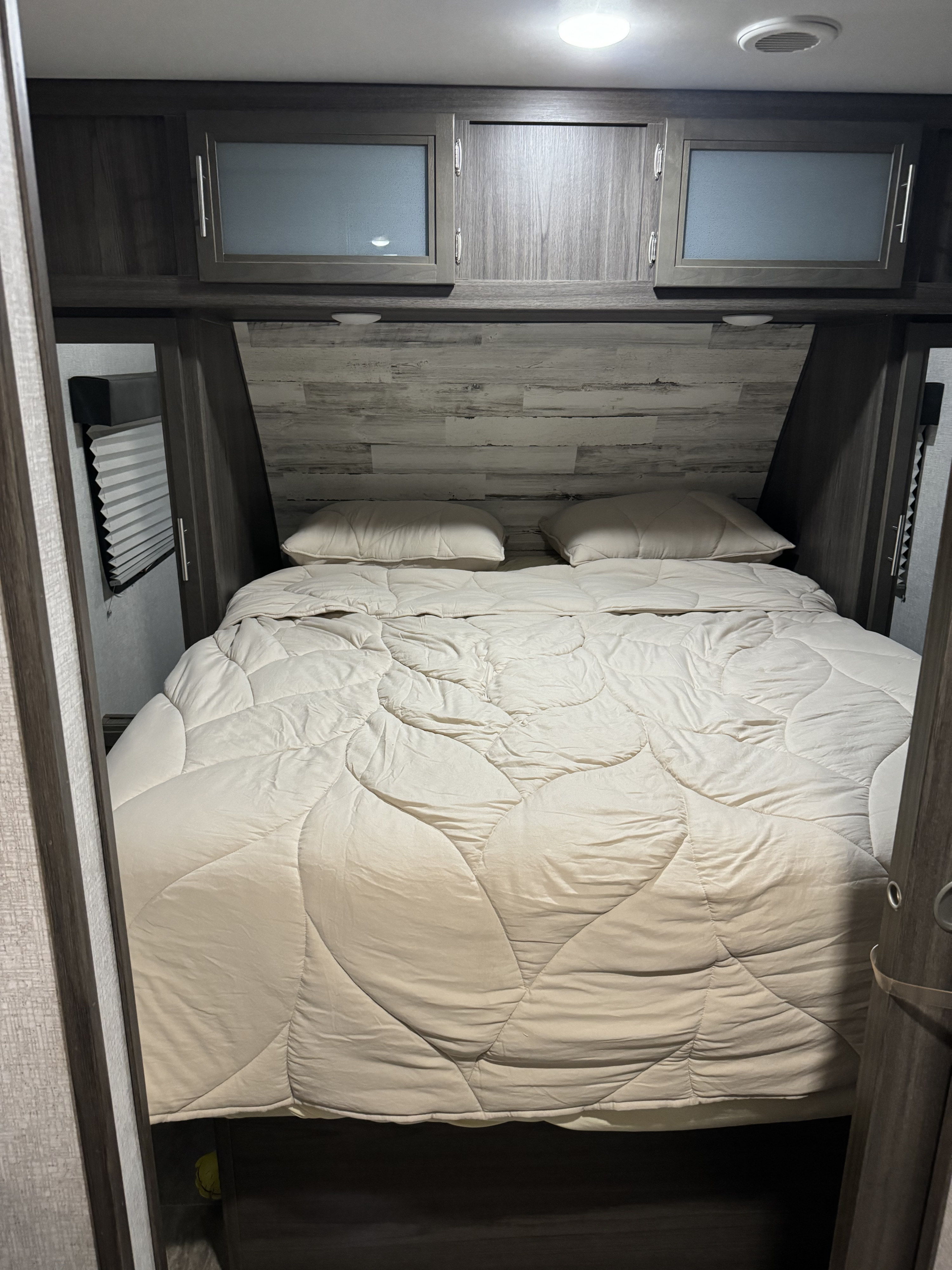 RV rental bedroom: a cozy bed with quilted comforter, overhead cabinets, ceiling lights, and wood-patterned walls.. Dutchmen Kodiak Ultra Lite 2020
