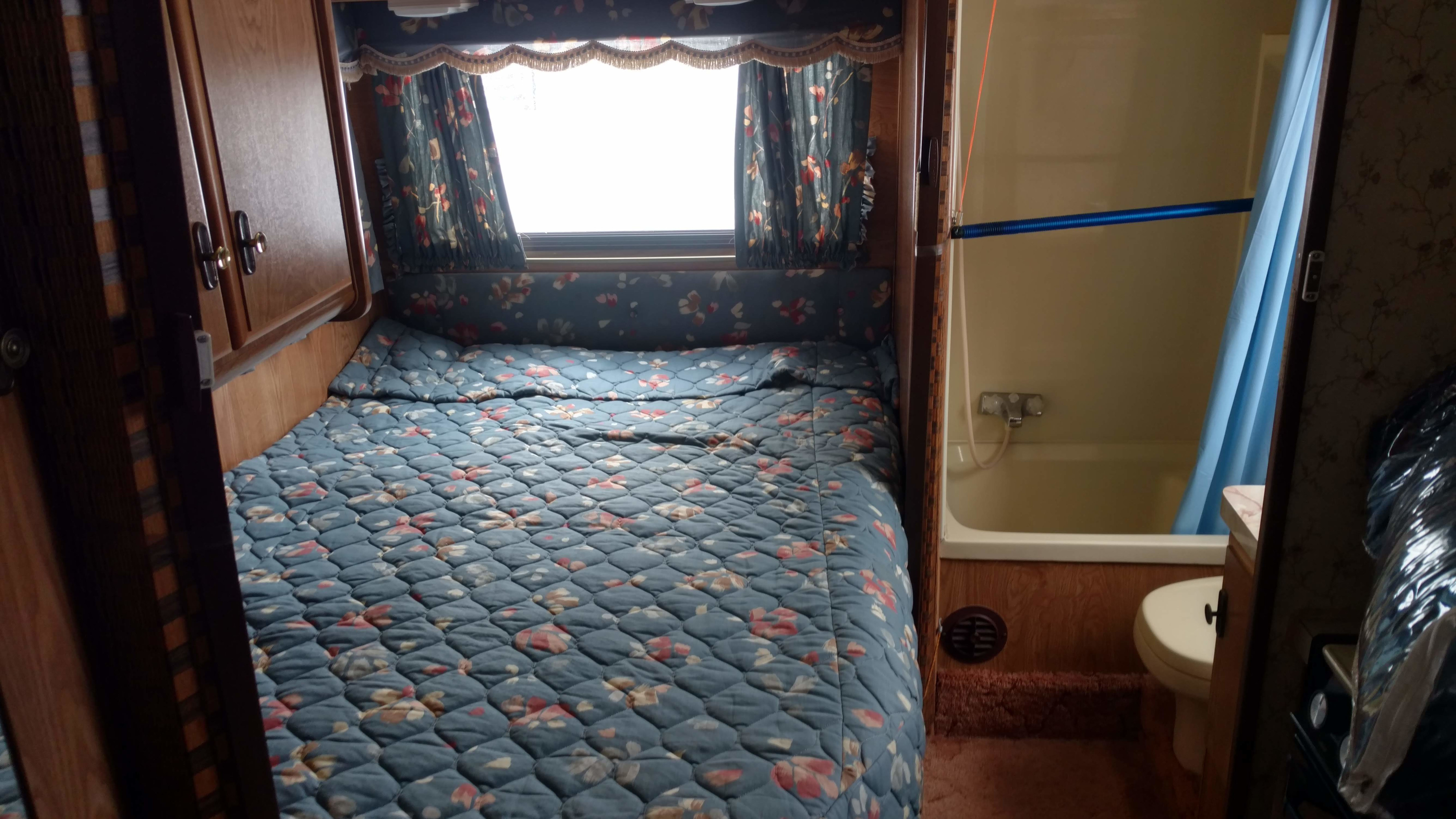Inside an **RV rental**: a snug bedroom with floral bedding, bright window, and an adjacent compact bathroom with tub/toilet.. Ford E350 Tioga Arrow 1988
