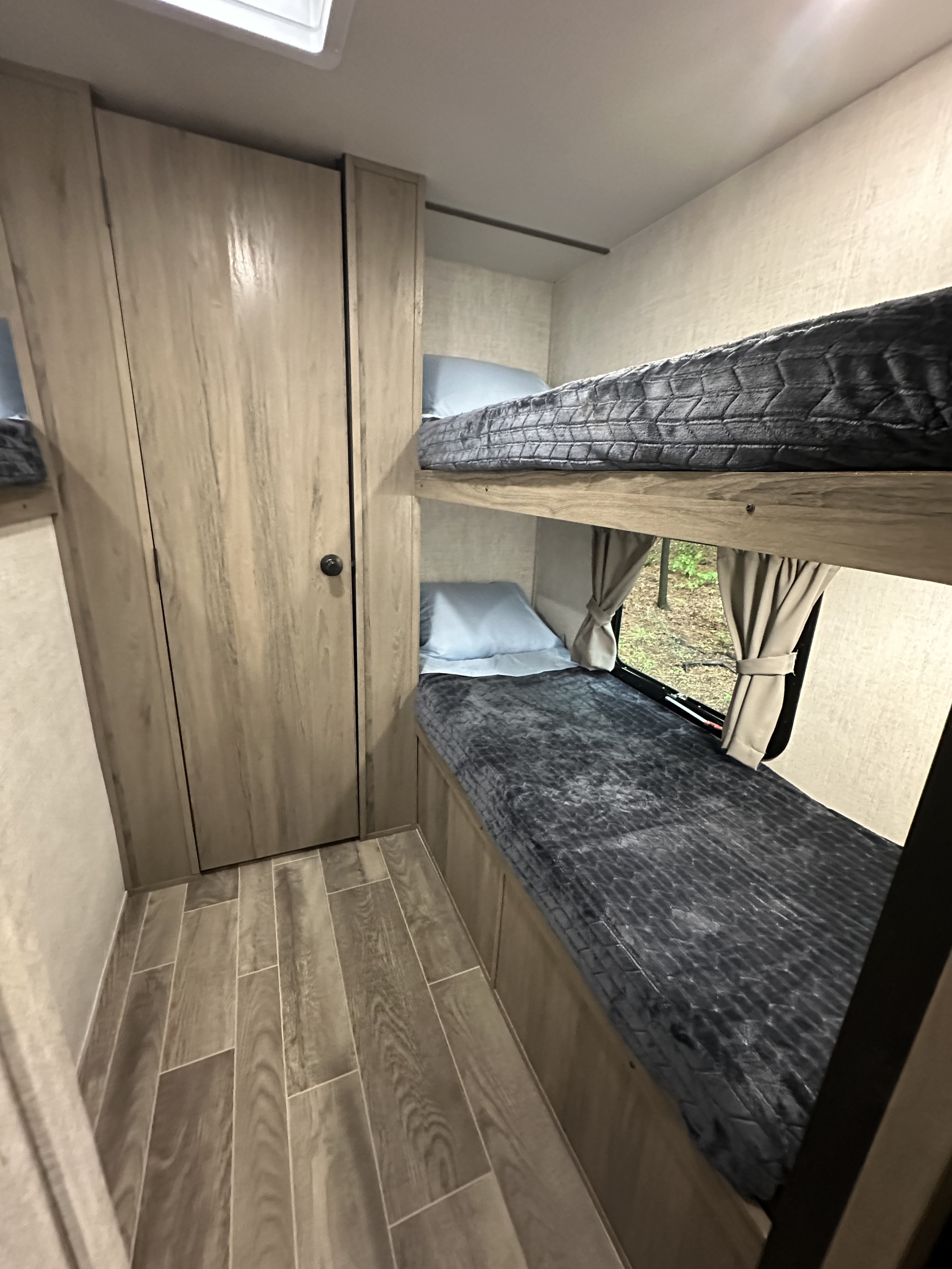 RV rental interior featuring cozy bunk beds with light pillows & dark blankets, wood-look cabinets, and plank flooring.. Gulf Stream Ameri-Lite 2021