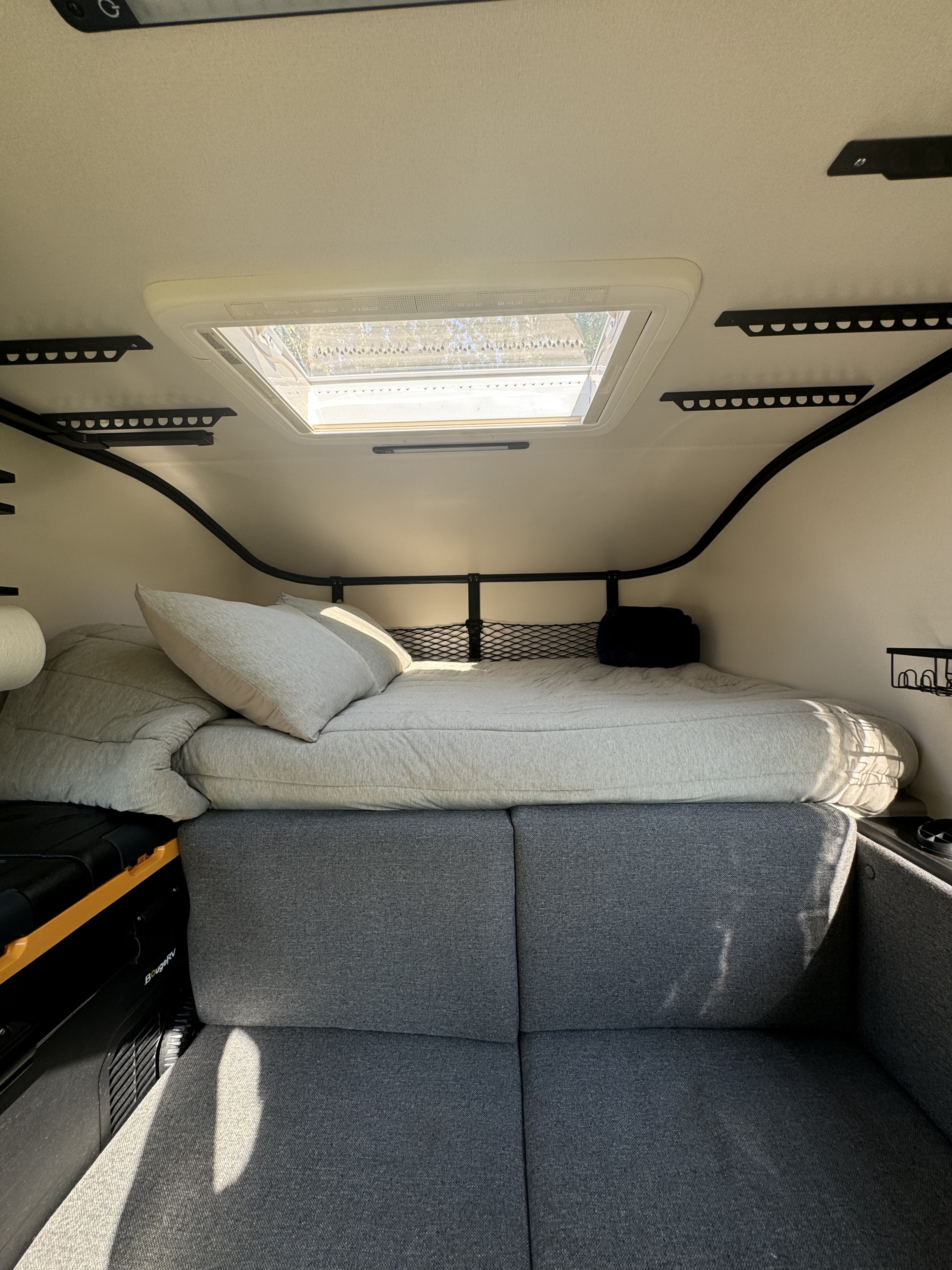 Bright RV rental interior with a cozy bed, gray modular couch, and a large skylight above.. Dodge 1500 2023